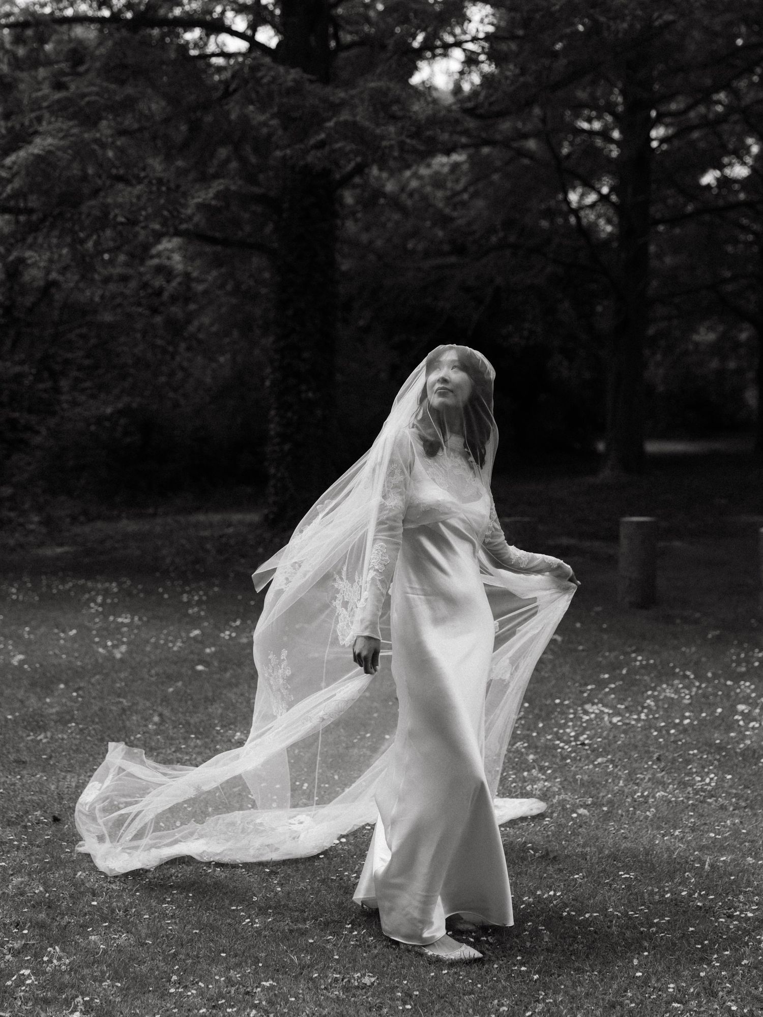 Black and white photo of a bride in a flowing gown and veil outdoors among trees.