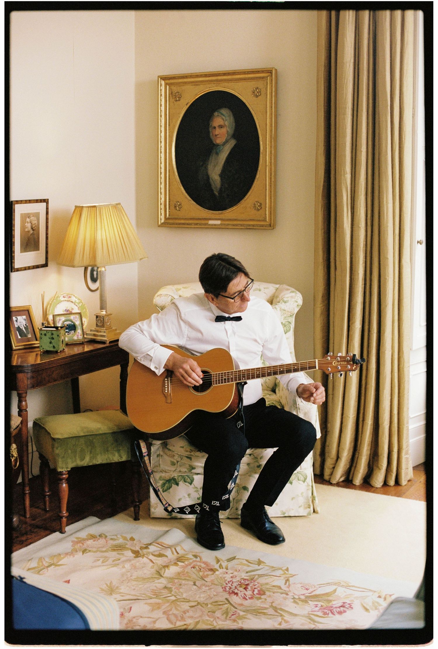 Young man in bow tie playing acoustic guitar in elegant room with portrait painting behind.
