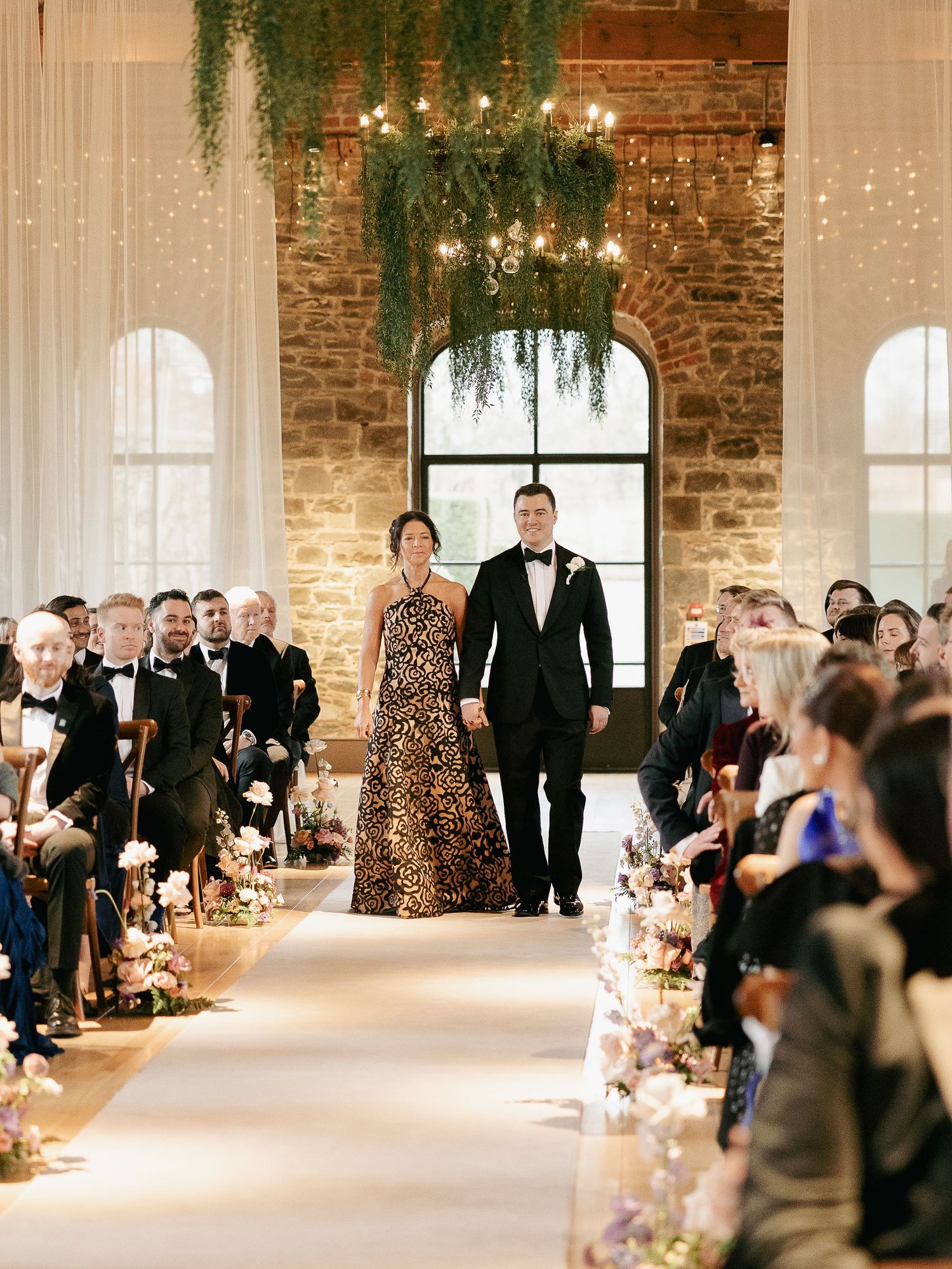 A wedding ceremony in a rustic brick venue with white drapes, hanging greenery, and candlelit aisle decorations.