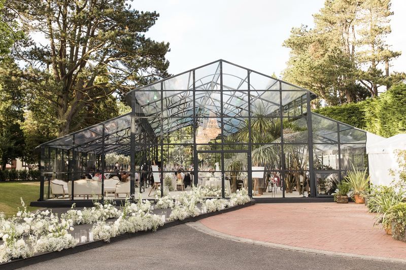 A panoramic view of a greenhouse walkway lined with white spring flowers and tall pine trees in a garden setting.