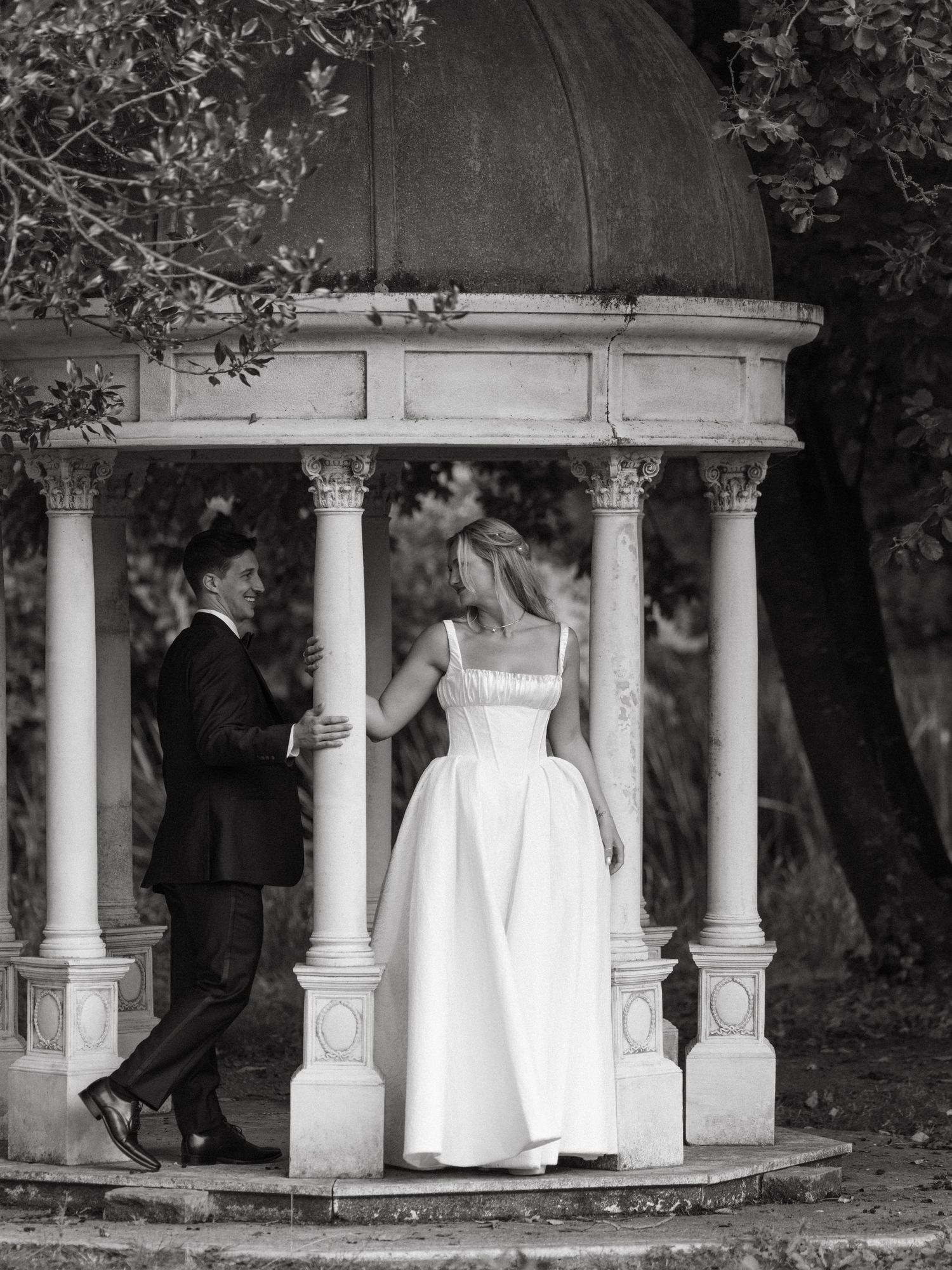 Black and white photo of a bride and groom sharing a romantic moment under an elegant stone gazebo.