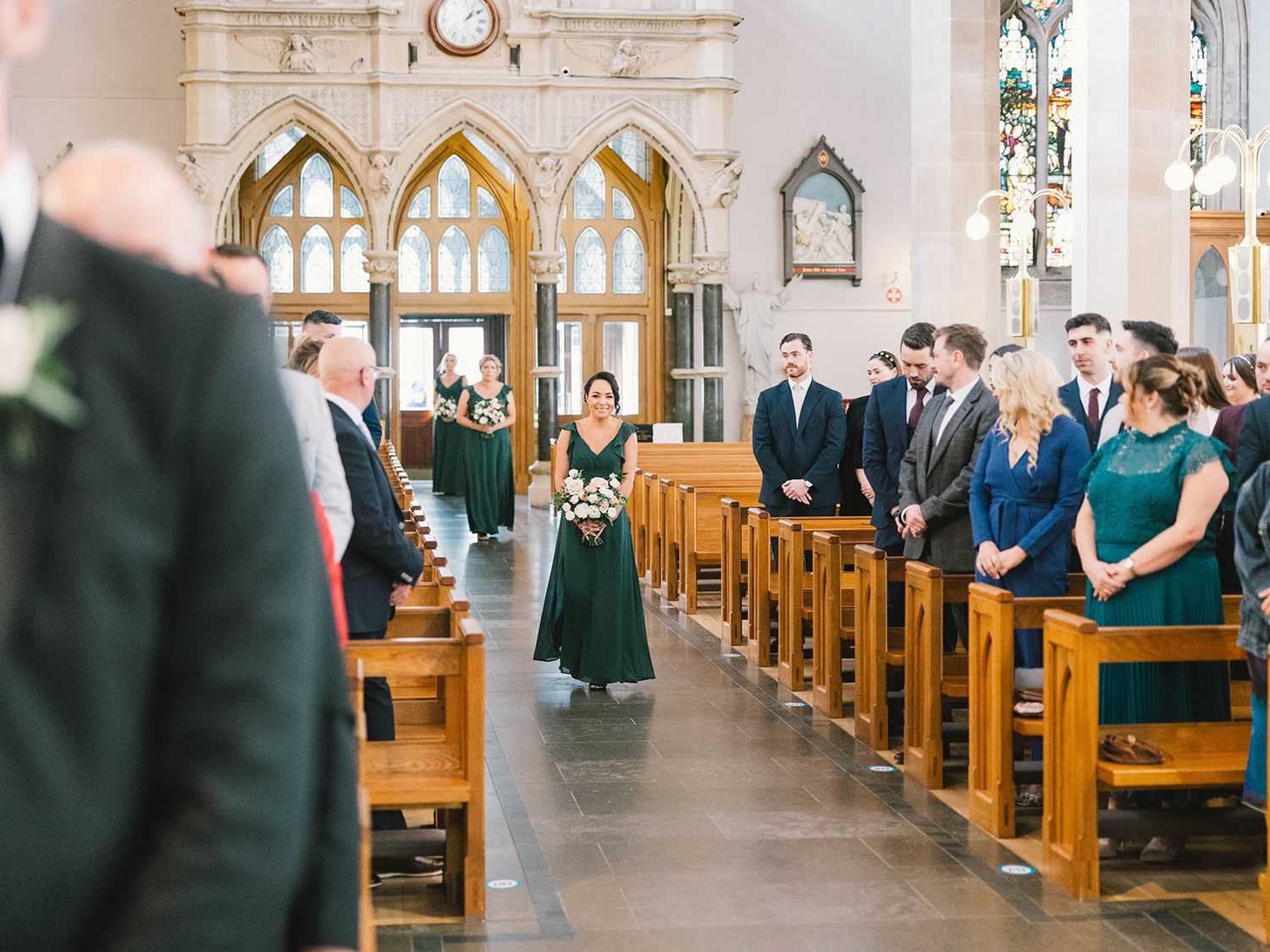 People stand in wooden pews during a ceremony inside an ornate gothic-style church with arched windows and white walls.