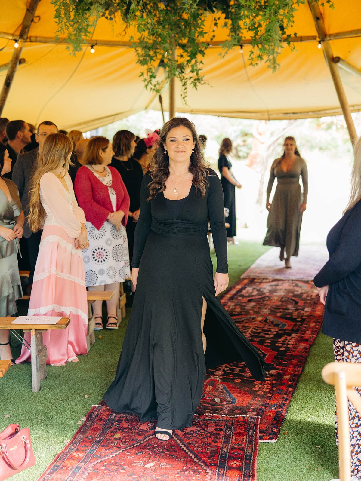 A guest in a black long dress walks down a red oriental rug under an outdoor tent at a formal event.