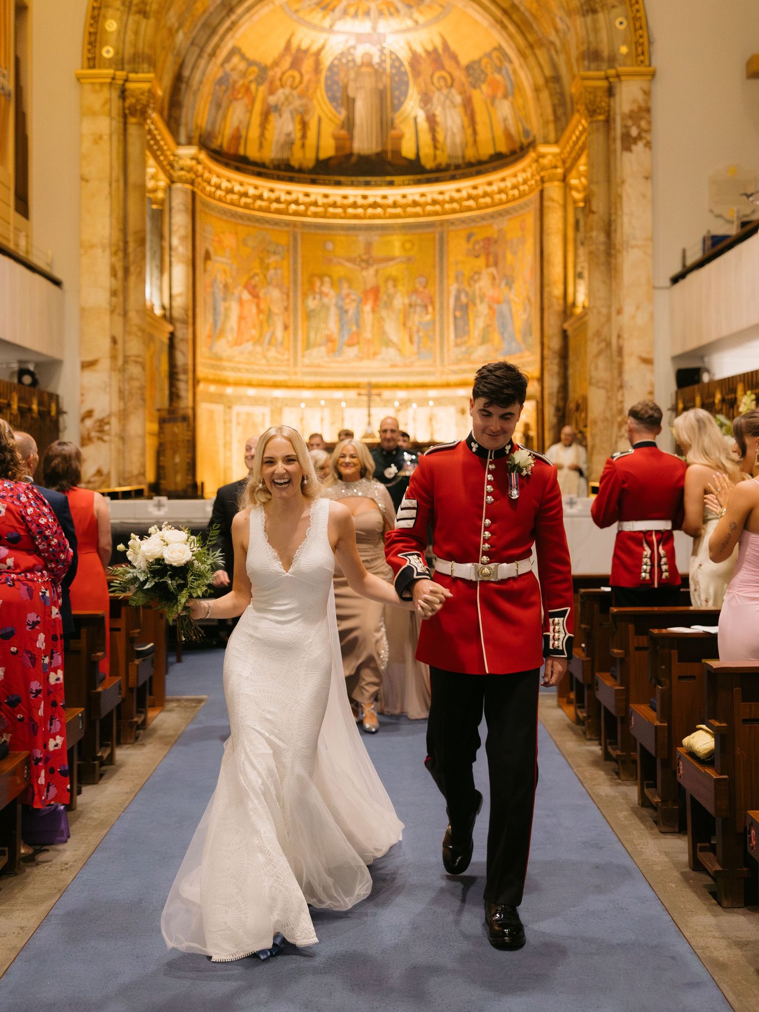 A wedding ceremony in a grand church with ornate golden apse mosaics as newlyweds walk down the blue-carpeted aisle.