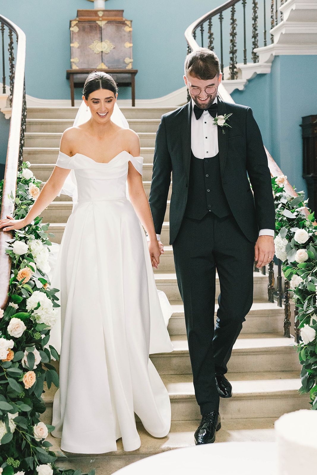 Elegant wedding couple descends grand staircase decorated with white floral arrangements and greenery.
