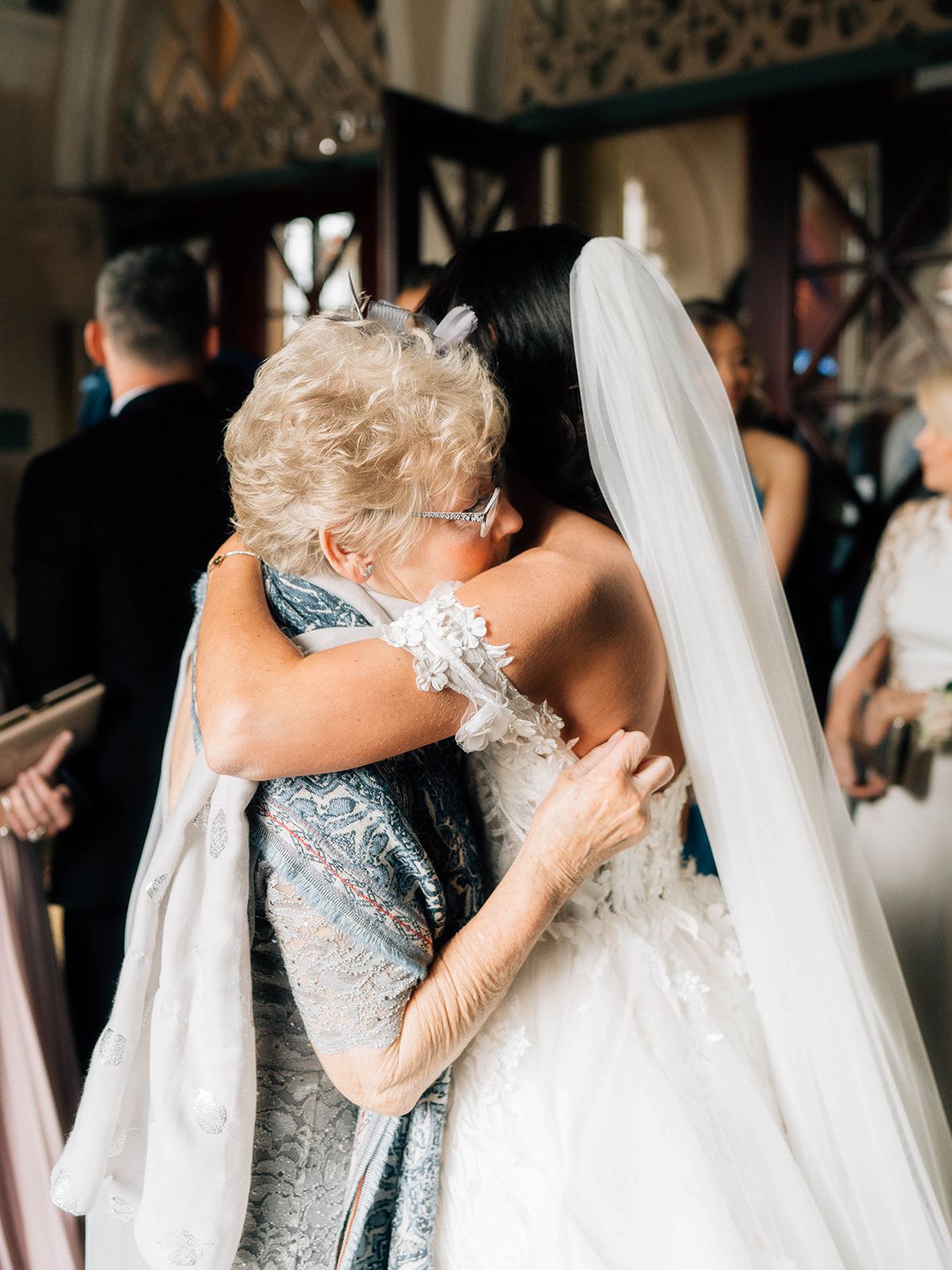 An emotional embrace between a bride in white and a guest wearing a blue dress at an elegant indoor wedding ceremony.