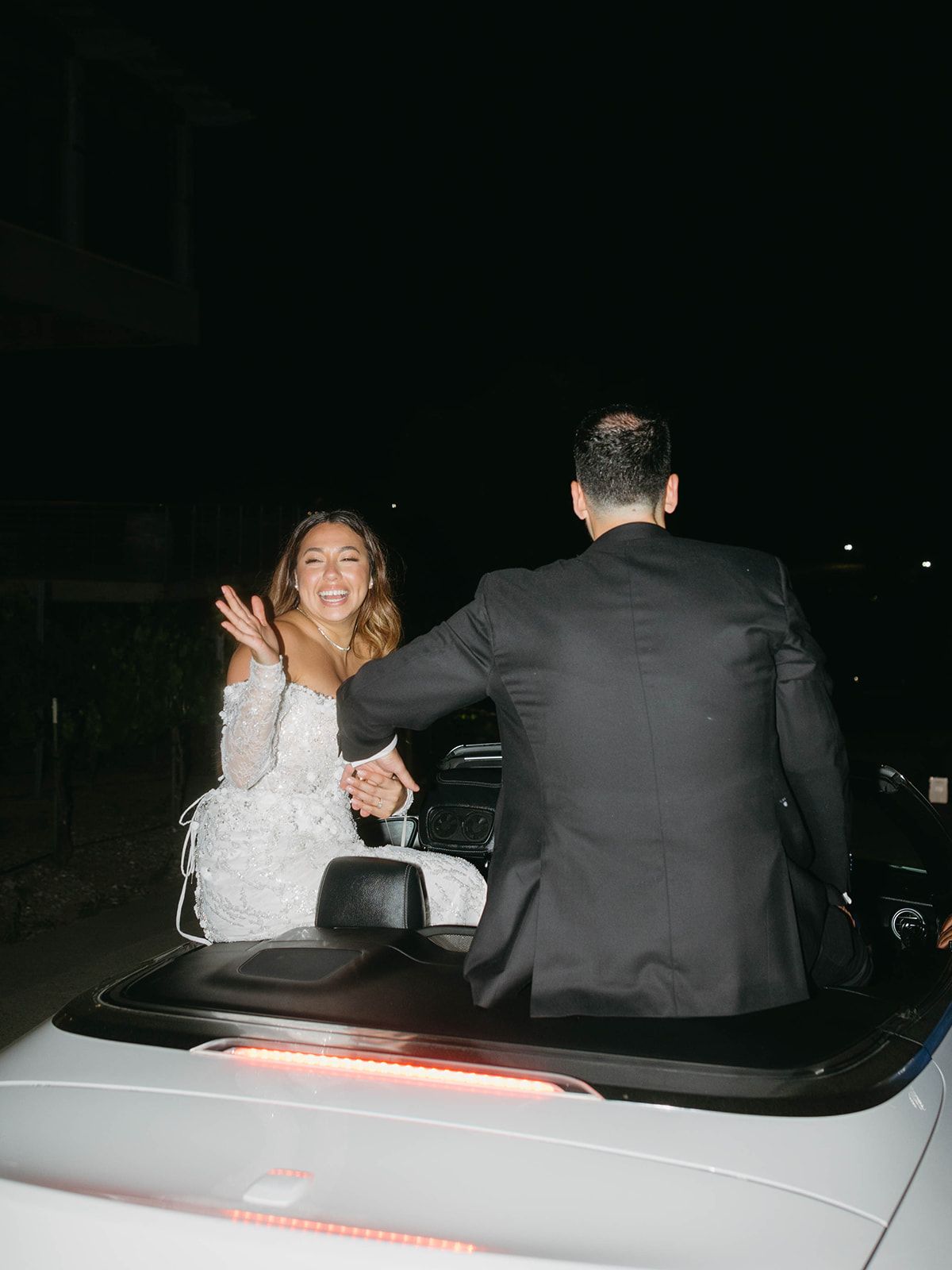 Bride and groom celebrating in the back of a classic white convertible car at night.