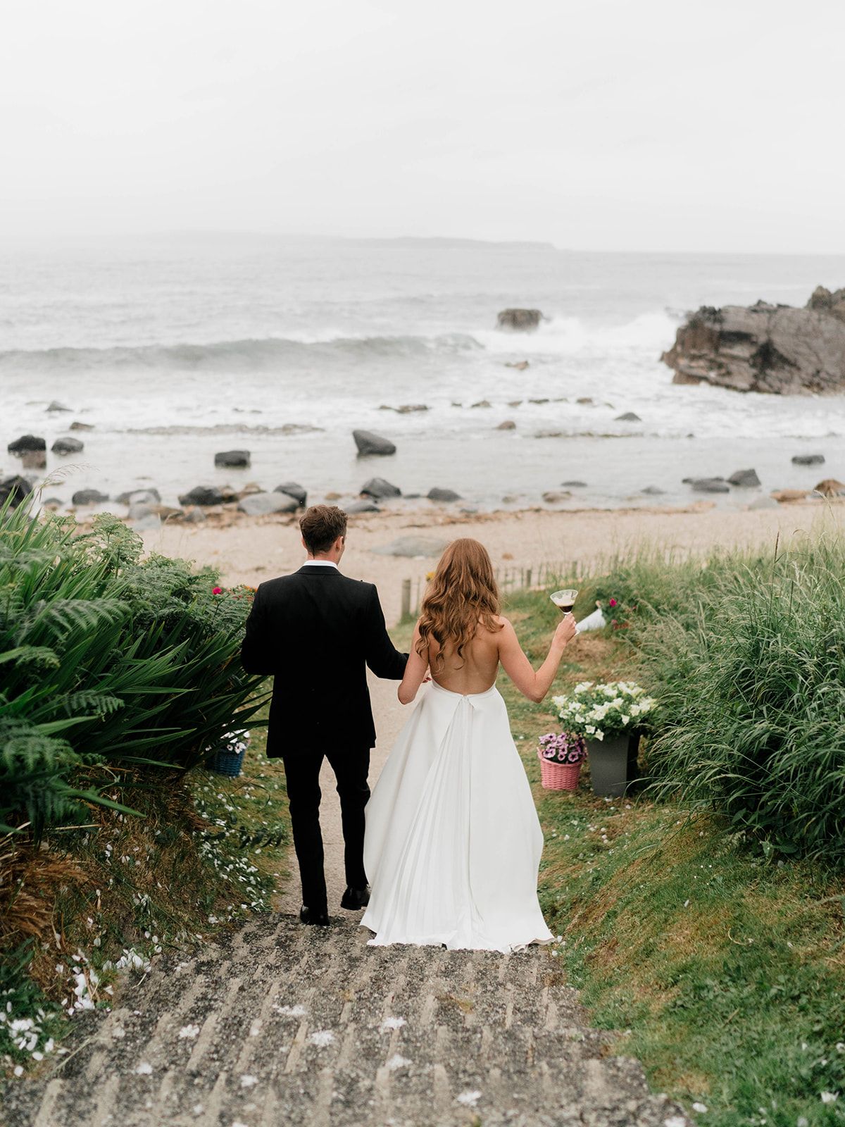 Couple walking down coastal path overlooking rocky beach on wedding day.
