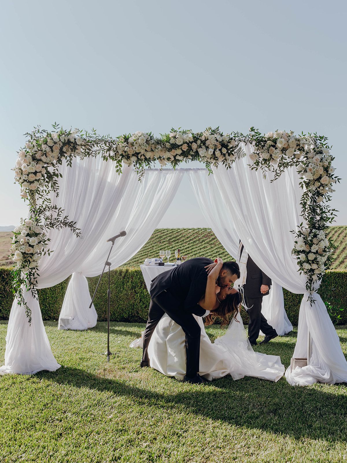 Wedding couple shares a moment under an elegant floral arch with flowing white drapes in a garden setting.
