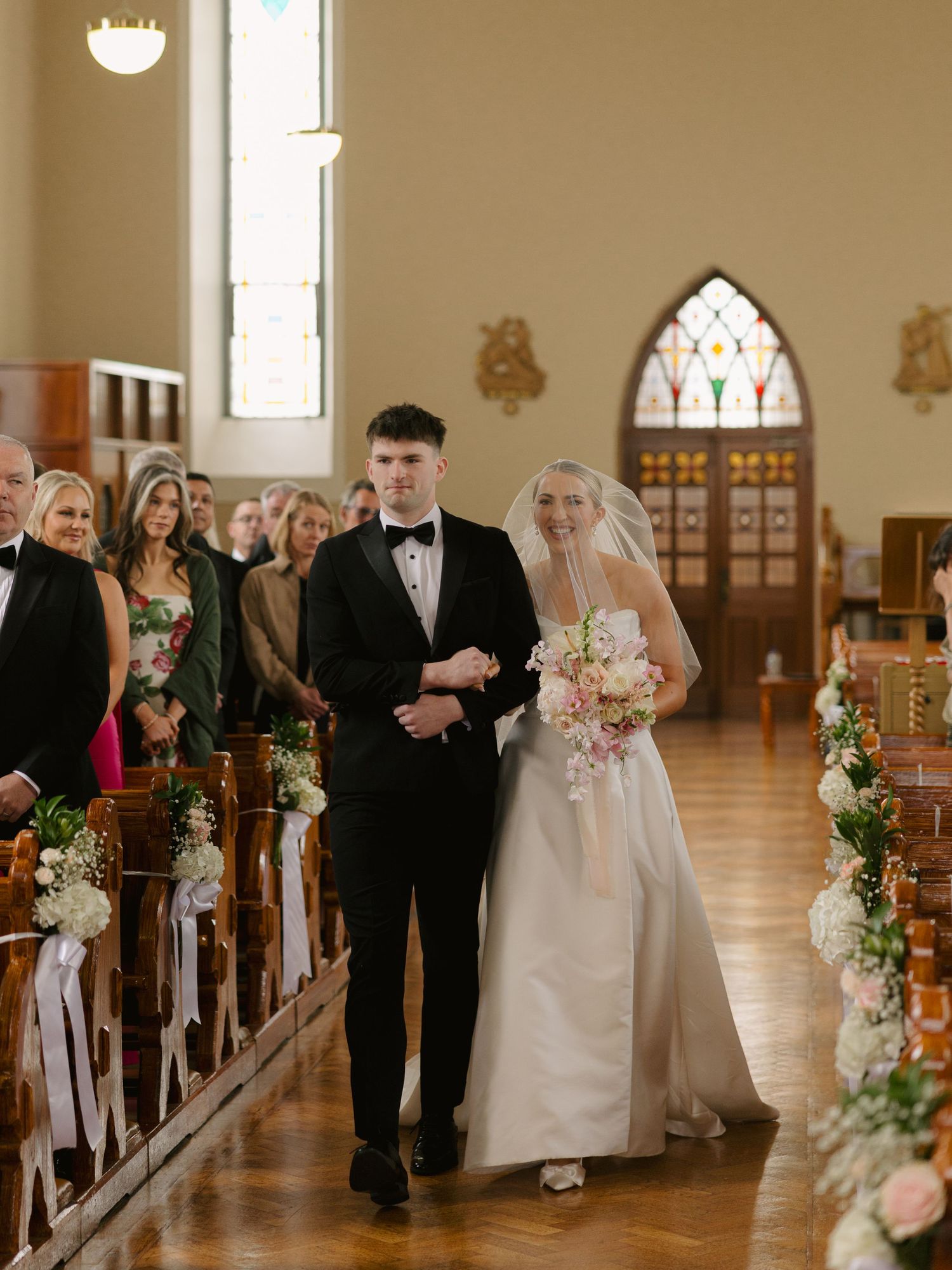 Bride and groom walk down church aisle during wedding ceremony, with floral arrangements and stained glass windows.
