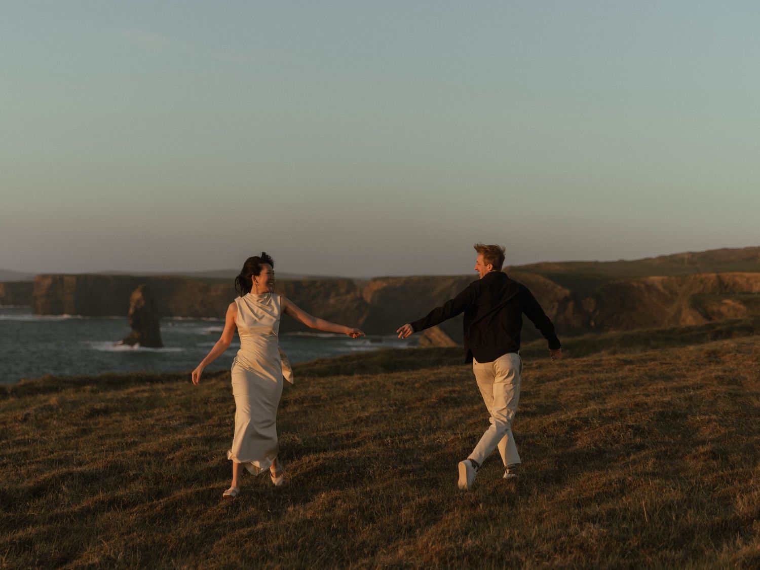 A couple running together on coastal cliffs during golden hour.