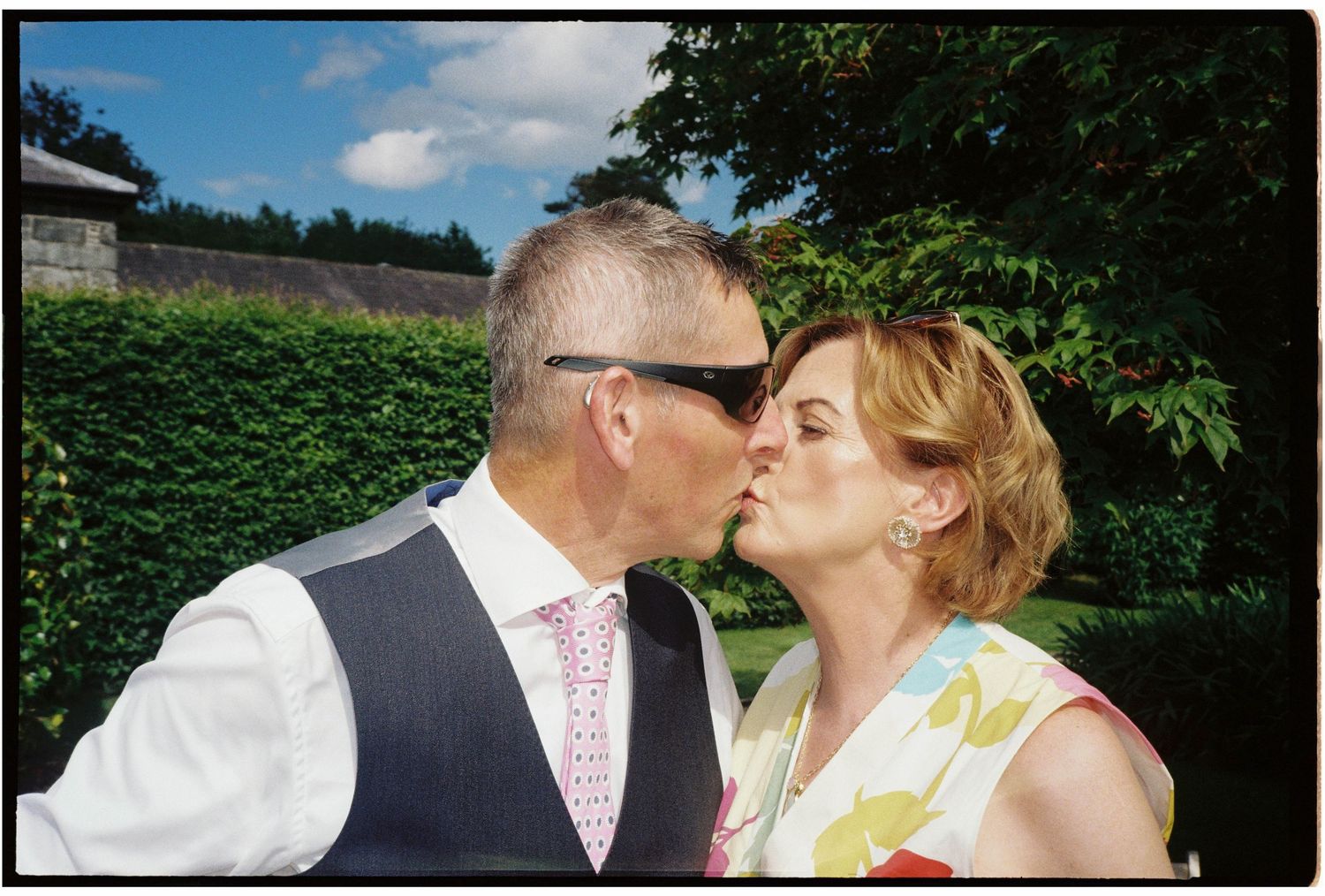 Couple sharing a romantic kiss outdoors at a wedding, man in vest and sunglasses, woman in floral dress.