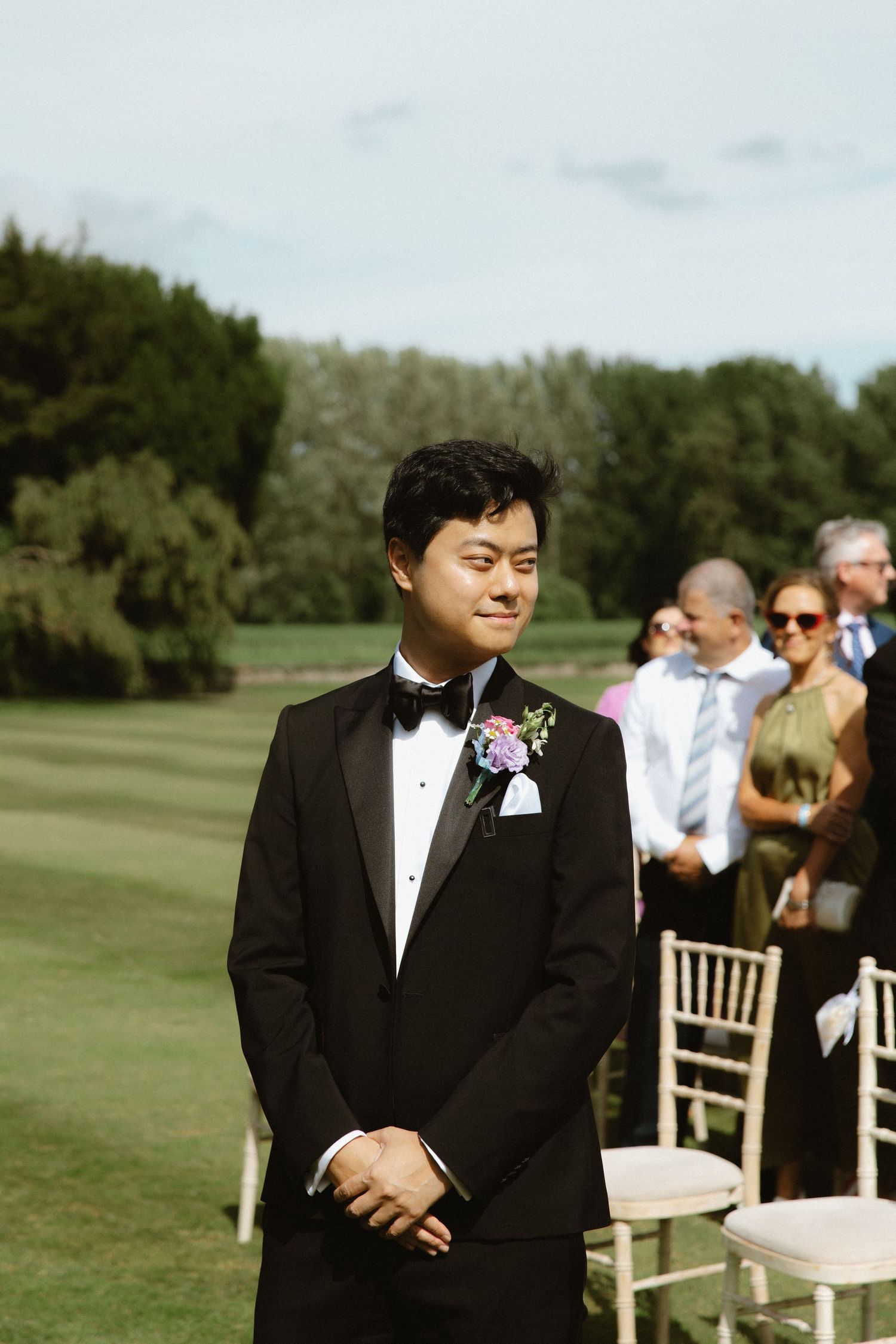 Groom in black tuxedo and purple boutonniere stands at outdoor wedding ceremony with guests in background.