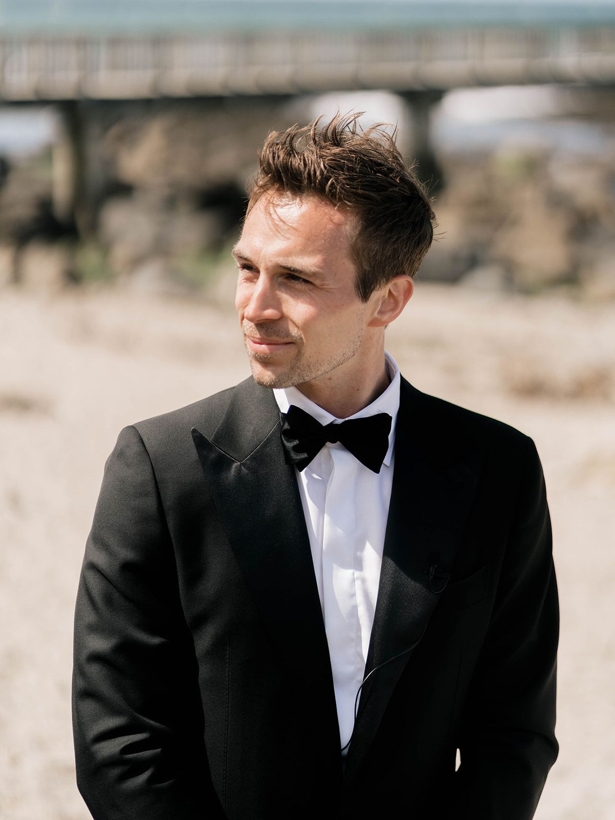 A groom in a classic black tuxedo and bowtie looks off to the side in an outdoor setting.