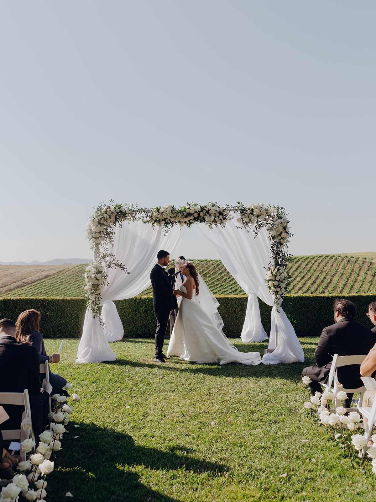 A wedding ceremony takes place under a floral arch with white drapes overlooking rolling vineyard hills on a sunny day.