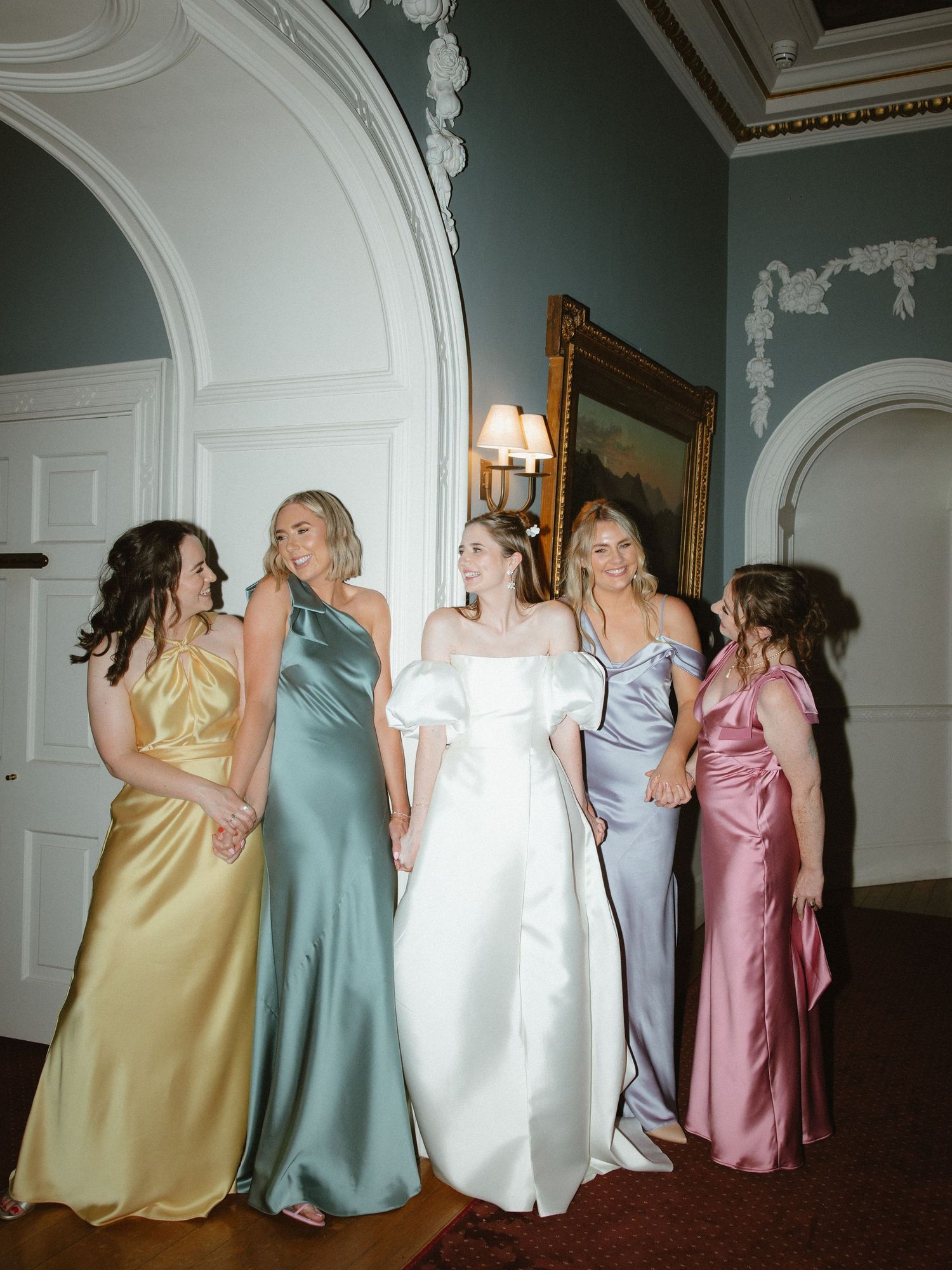 Bridesmaids in pastel silk dresses pose together in an elegant room with ornate moldings.