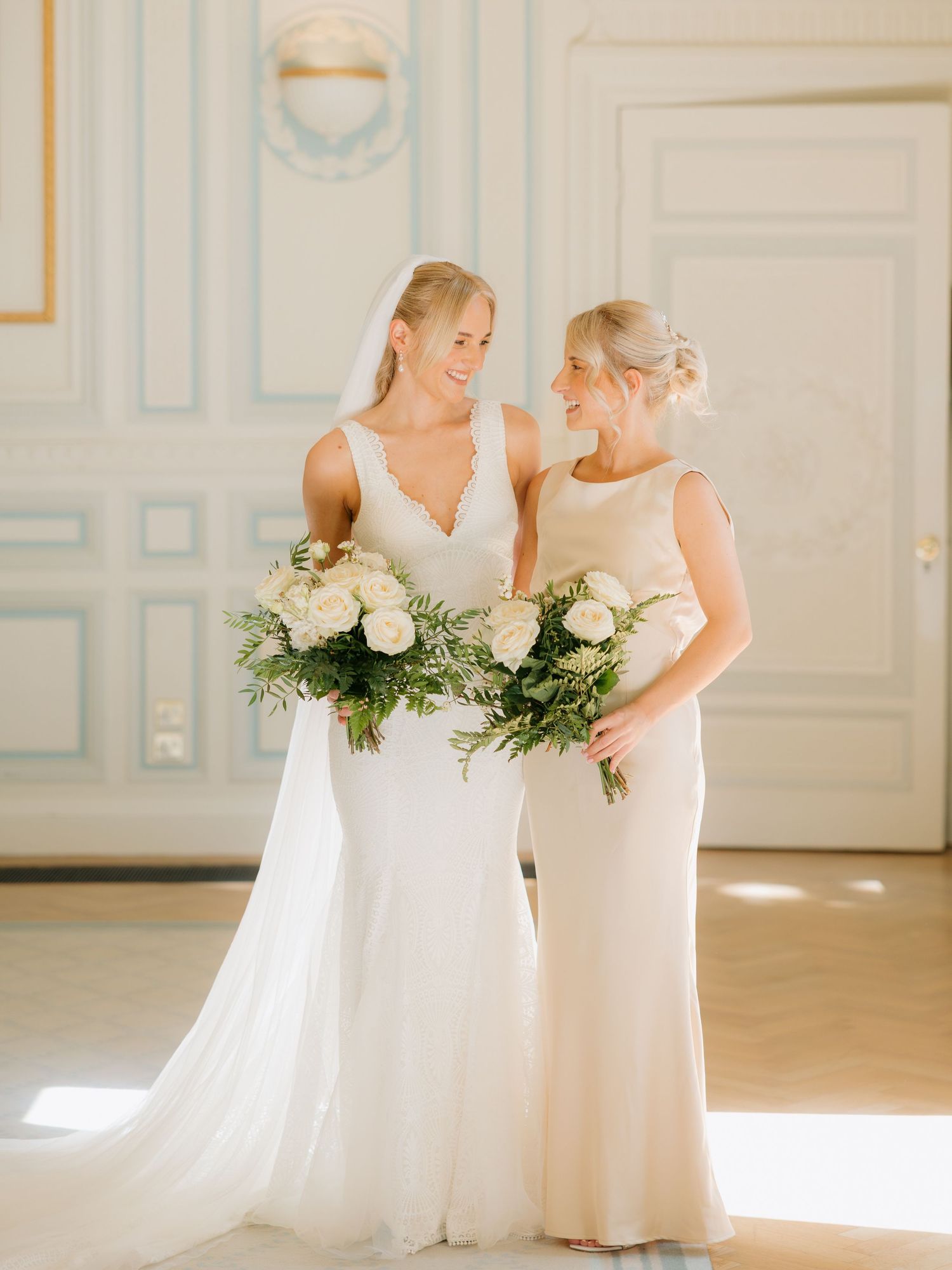 Bridesmaids in elegant white dresses pose playfully in a bright sunlit room with ornate wall details.