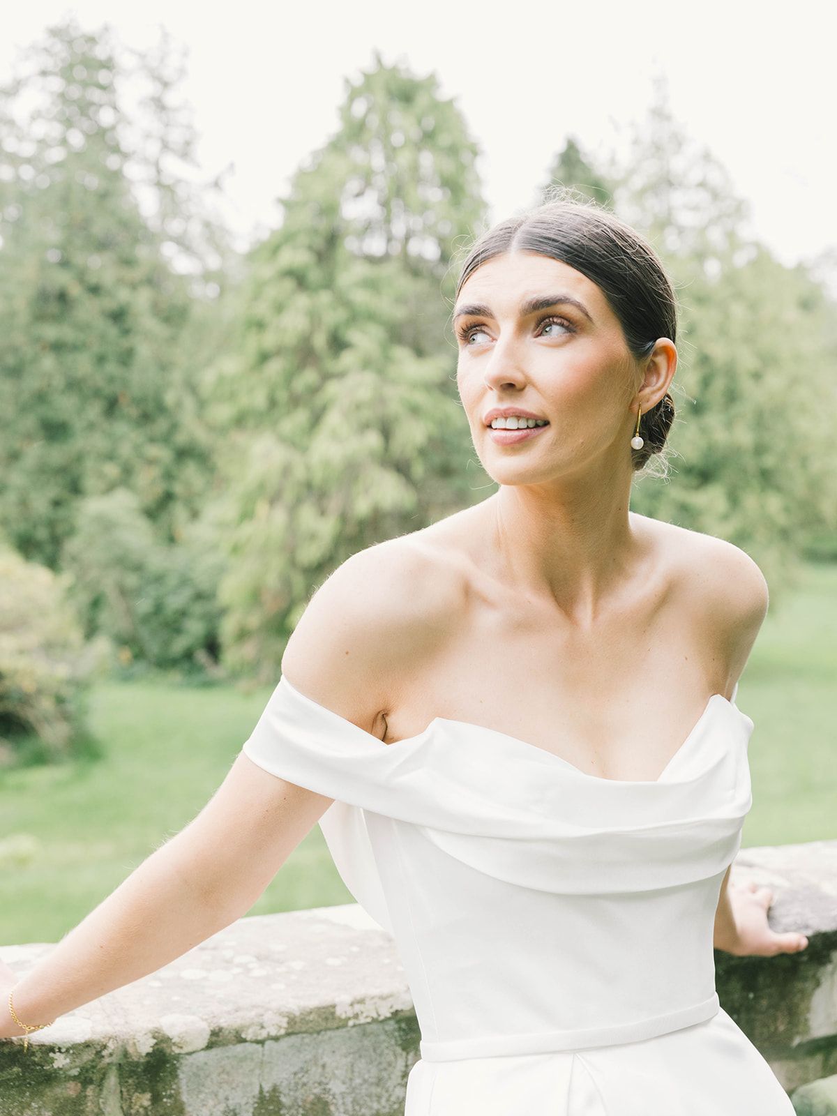 Woman in elegant white off-shoulder wedding dress smiles while looking up in outdoor garden setting.