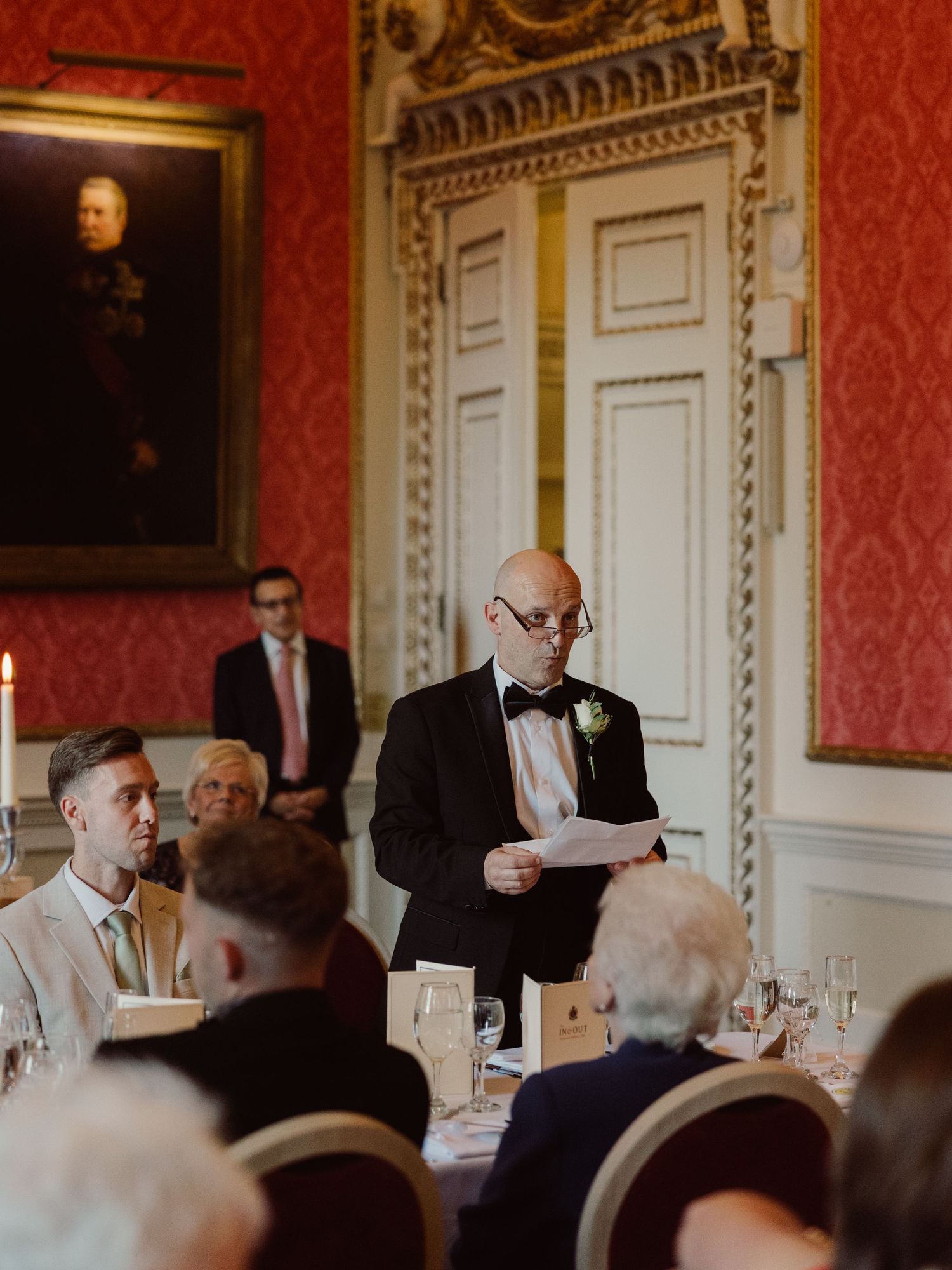 Guests seated at formal dinner tables with ornate decor and red damask wallpaper in an elegant historical room.