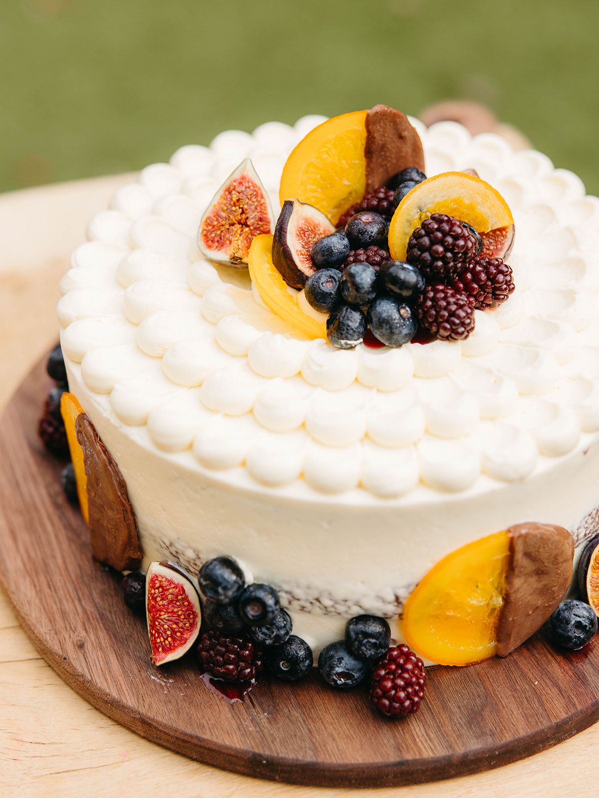 White frosted cake decorated with fresh fruits including figs, blackberries, blueberries, and orange slices on a wooden board.
