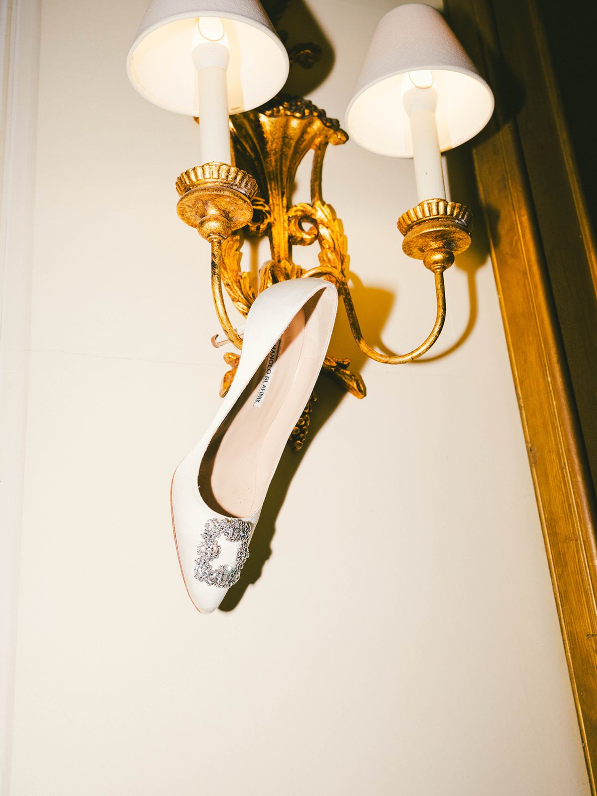 Gold ornate wall sconce with white shades illuminates a silver sequined ballet flat hanging on display.