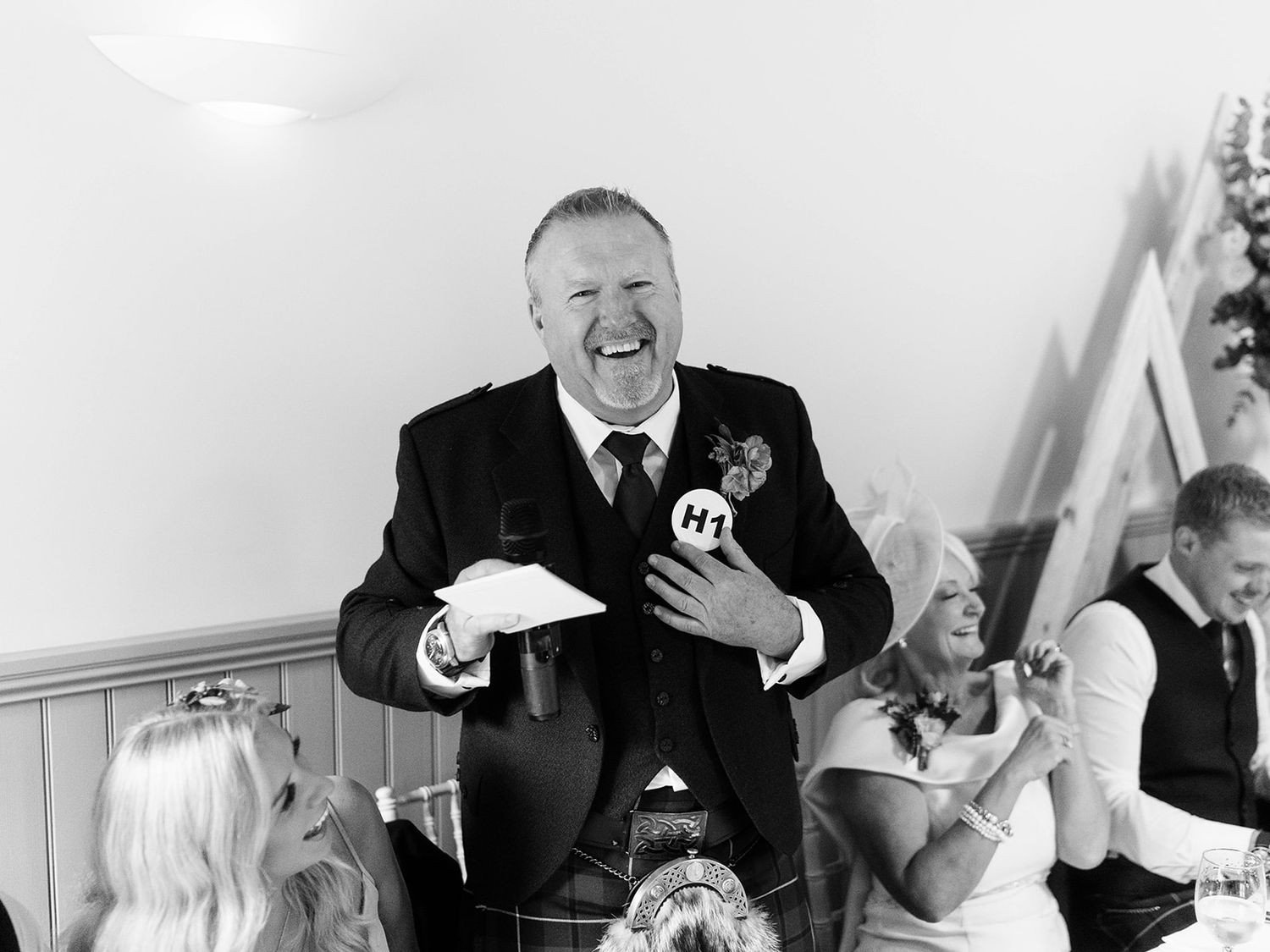 A person in a black suit laughs heartily during what appears to be a wedding reception speech or toast.