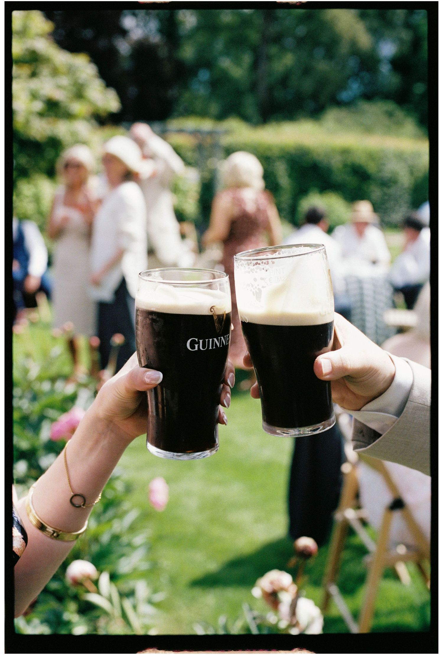 Two hands clink Guinness pint glasses in a sunny garden wedding reception with guests mingling in the background.