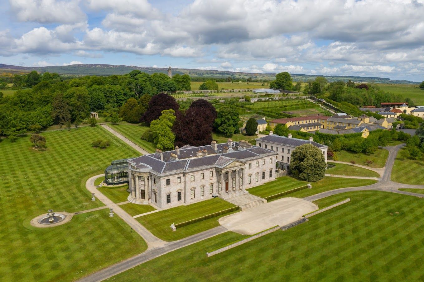 Aerial panoramic view of a grand manor house with manicured lawns and gardens in the countryside.