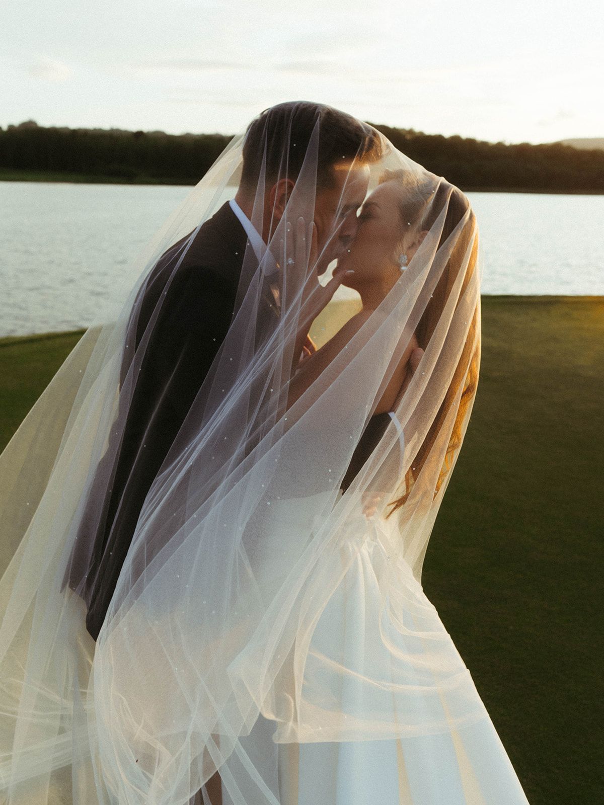 A romantic wedding couple embraces under a flowing veil at sunset by a lake on a grassy shore.