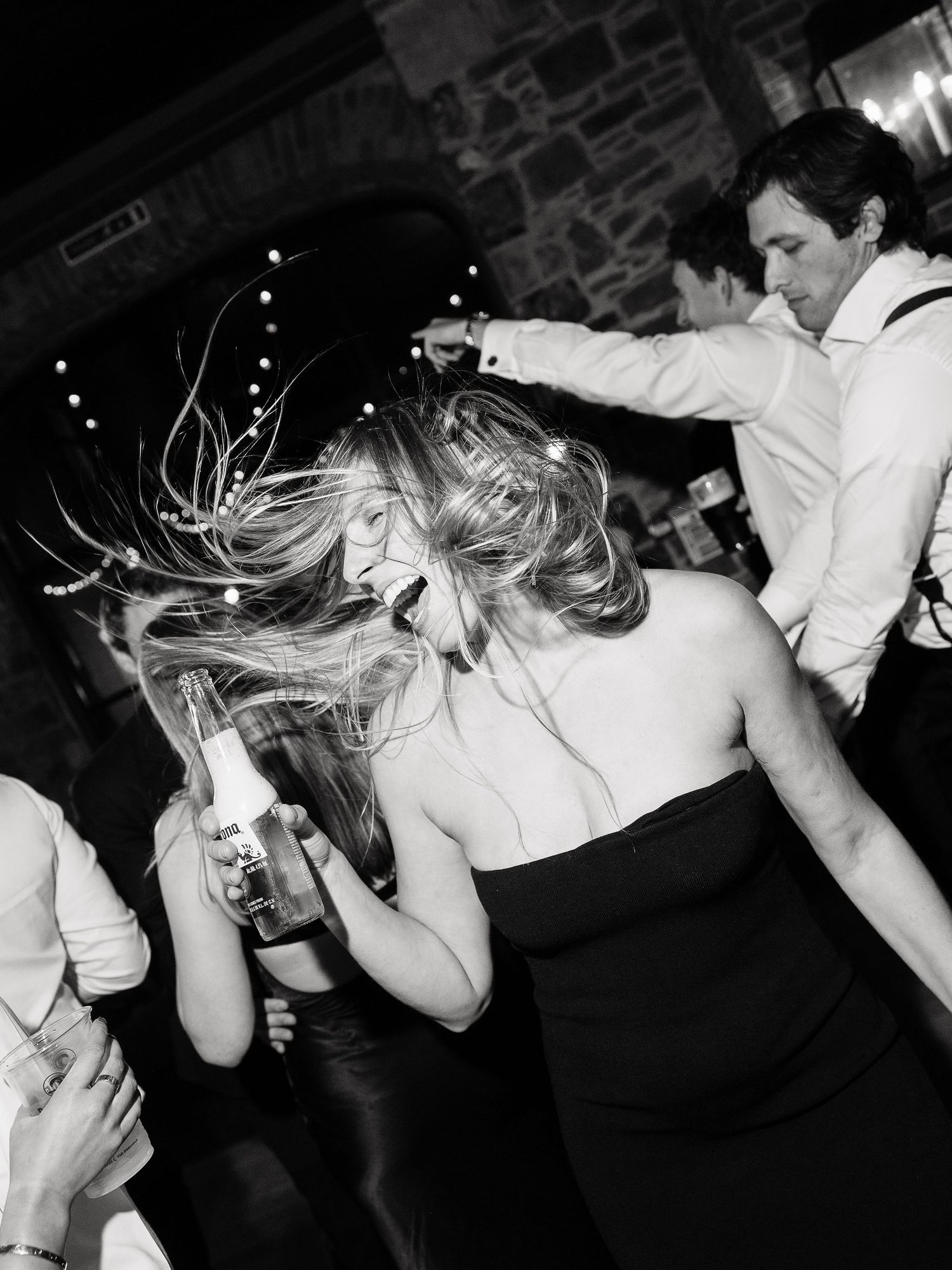Black and white photo of energetic dancing at a party with hair flying and drinks in hand on a crowded dance floor.