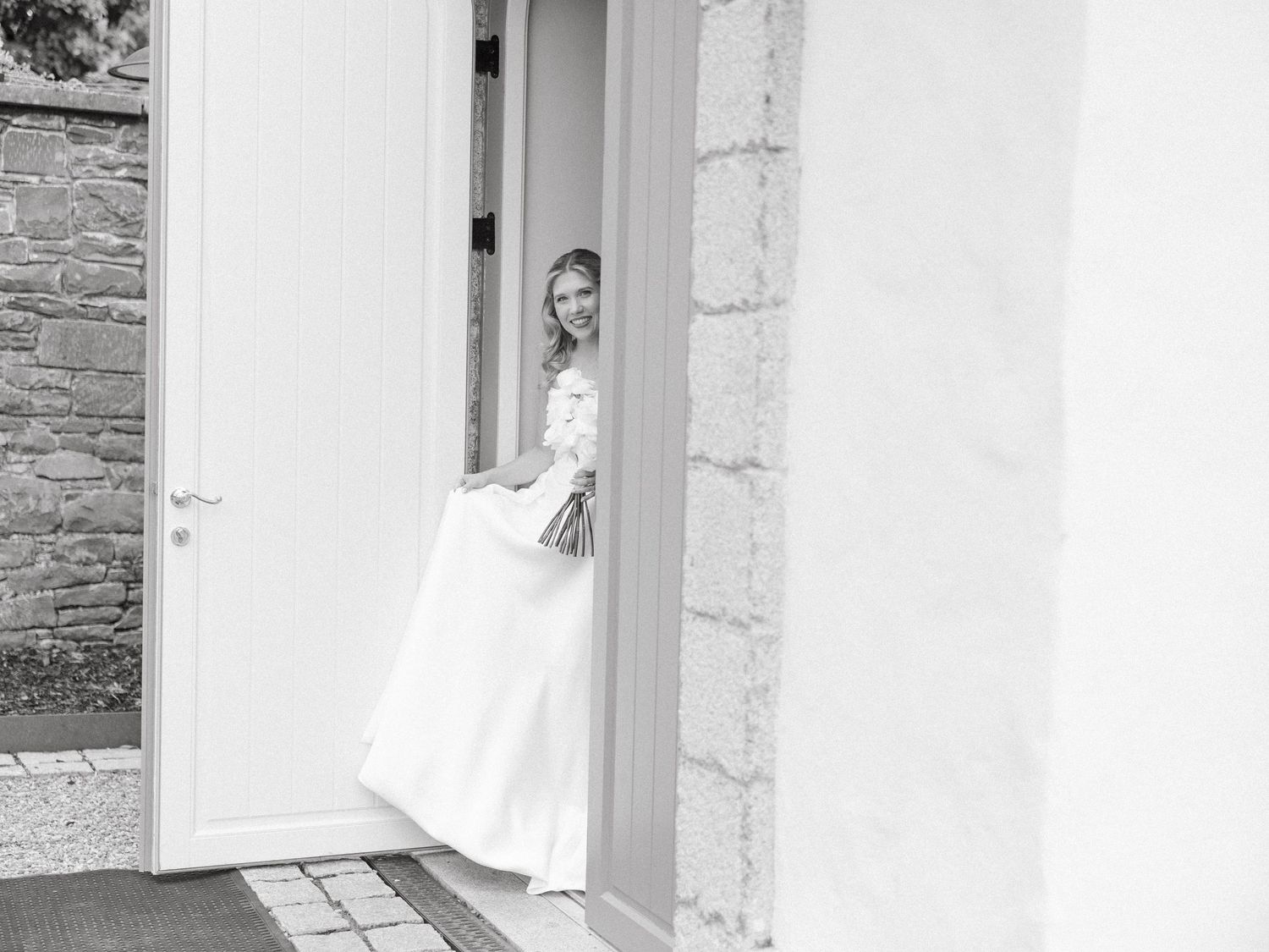 A person in a white dress peeks out from behind a doorway in a black and white photograph.