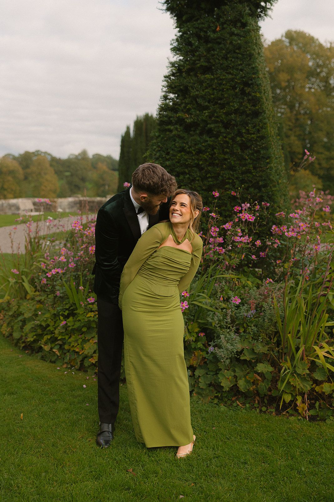 Bridesmaids in olive green dresses pose playfully in a garden with blooming pink flowers.
