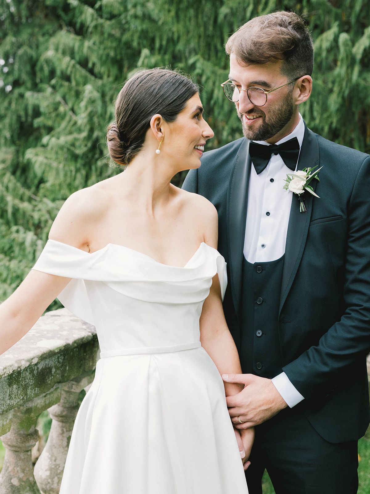 Newlywed couple in formal wedding attire smiling at each other against lush greenery backdrop.