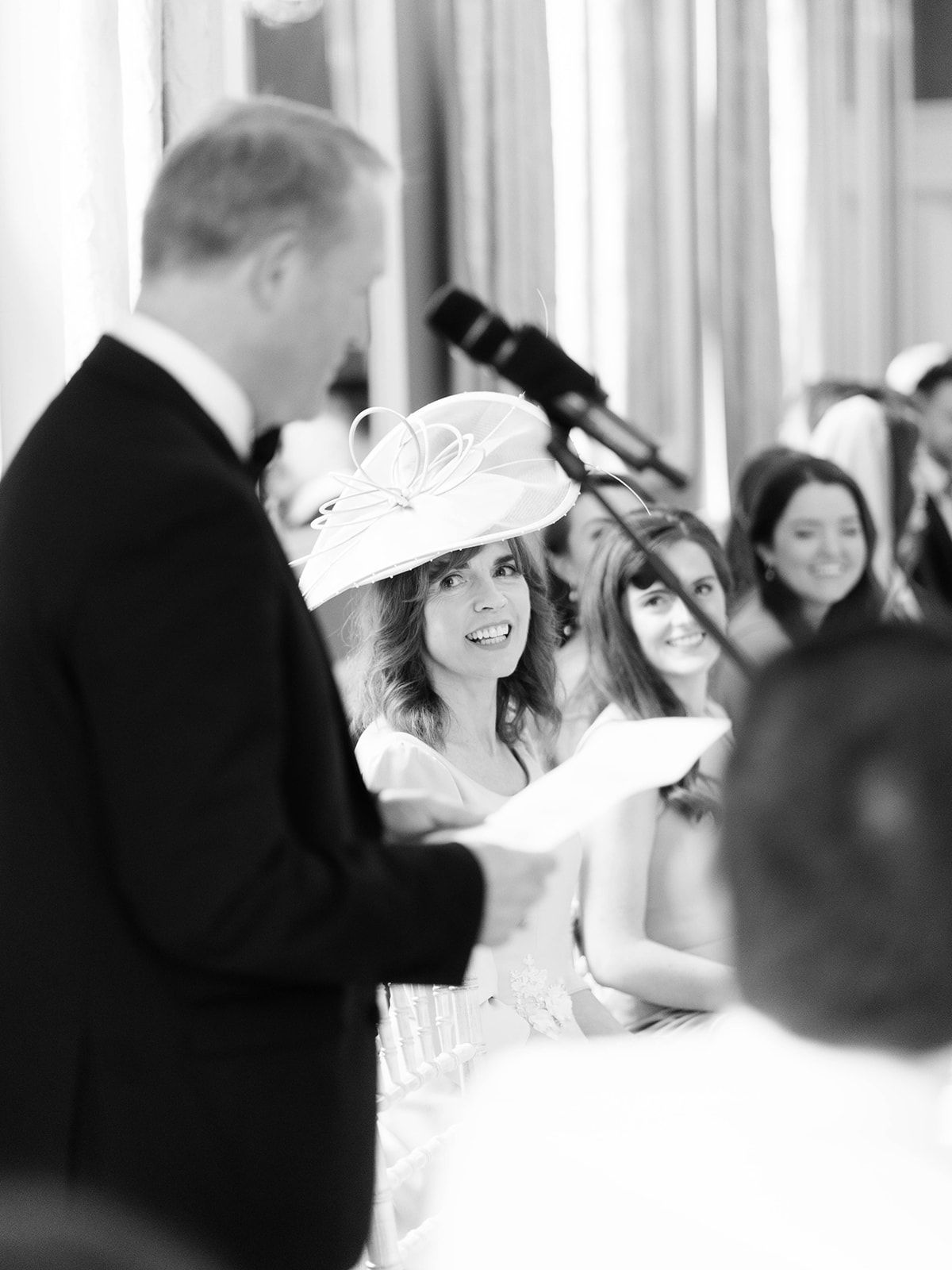 A bride in a white dress and decorative hat looks up during her wedding ceremony while standing by the microphone.