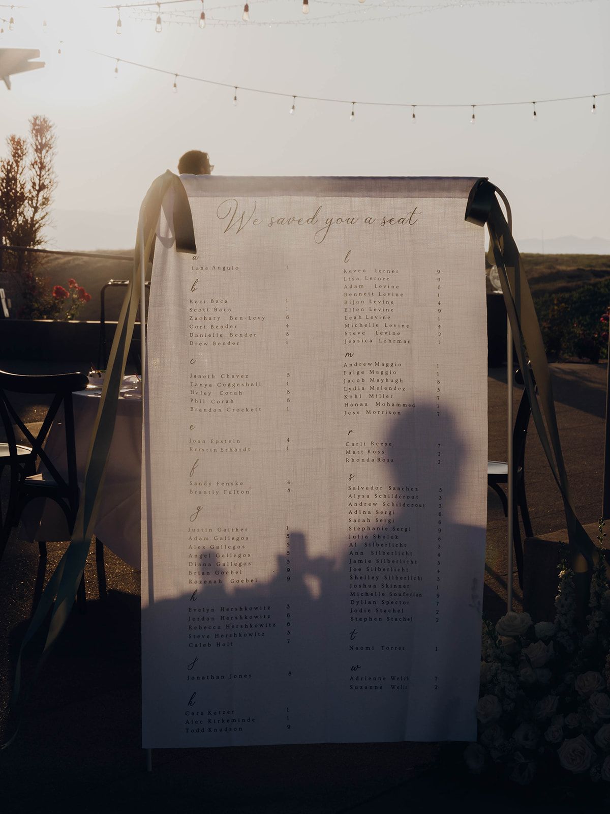 A wedding seating chart display with string lights hanging above during golden hour sunset.