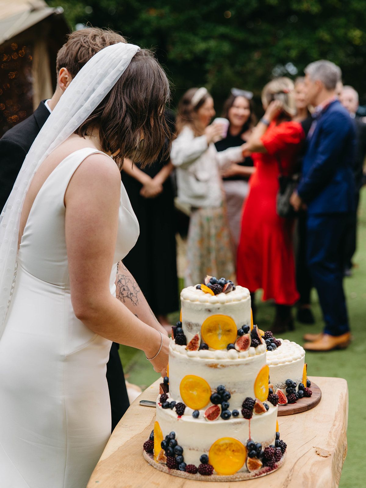 Wedding cake decorated with oranges and berries on display at outdoor reception with guests mingling.