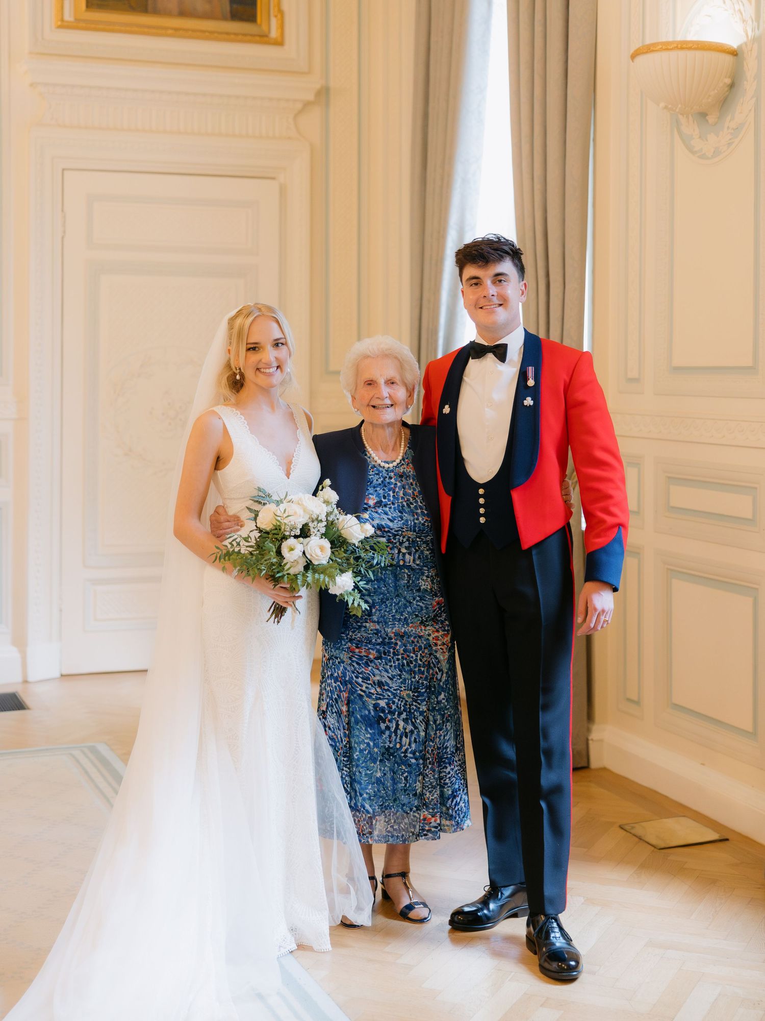 Three people pose formally in an elegant interior with white paneled walls, including a bride in white and a person in red military dress.