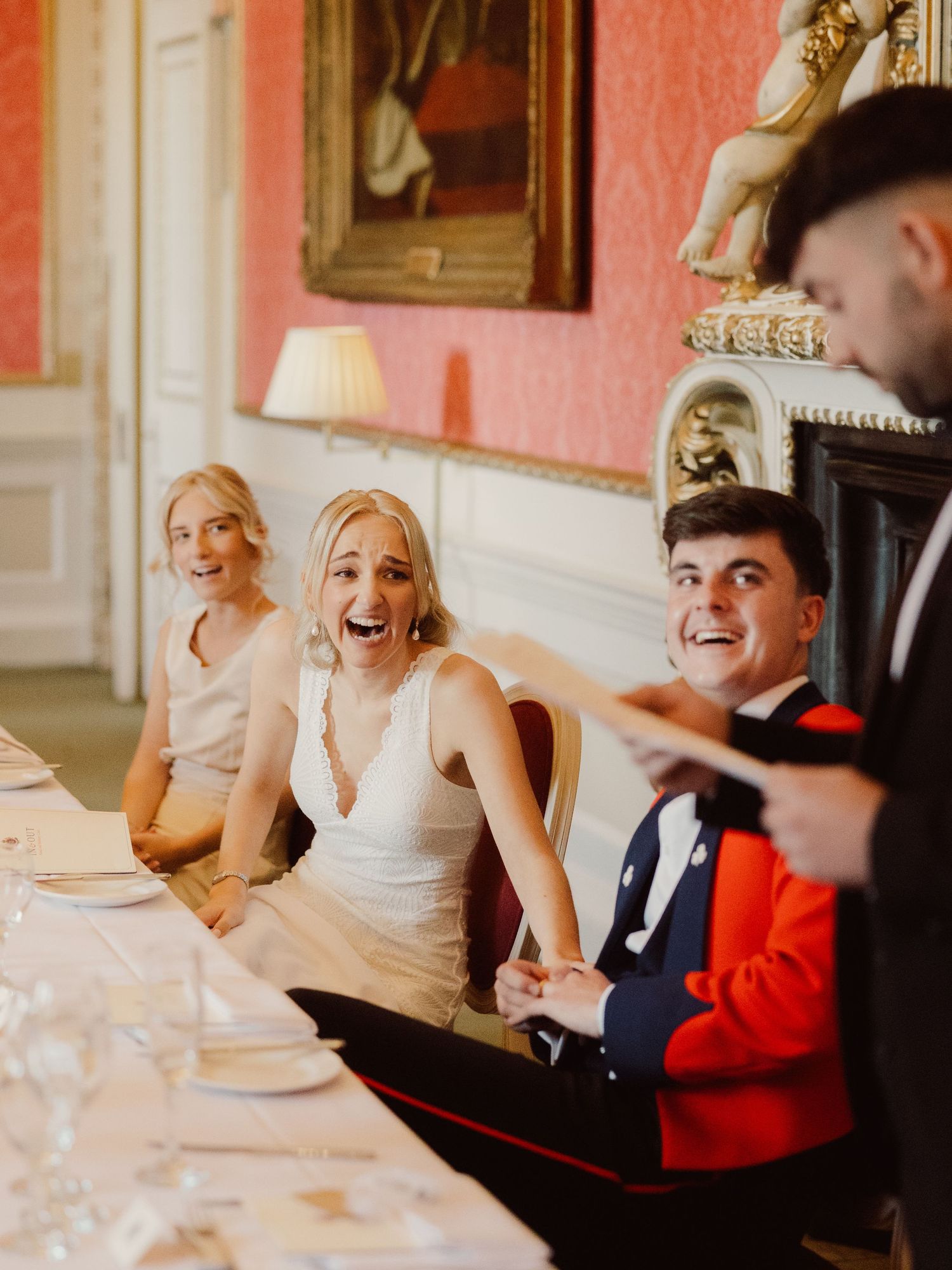 Wedding party members laughing together while seated on an elegant red couch in a formal room setting.