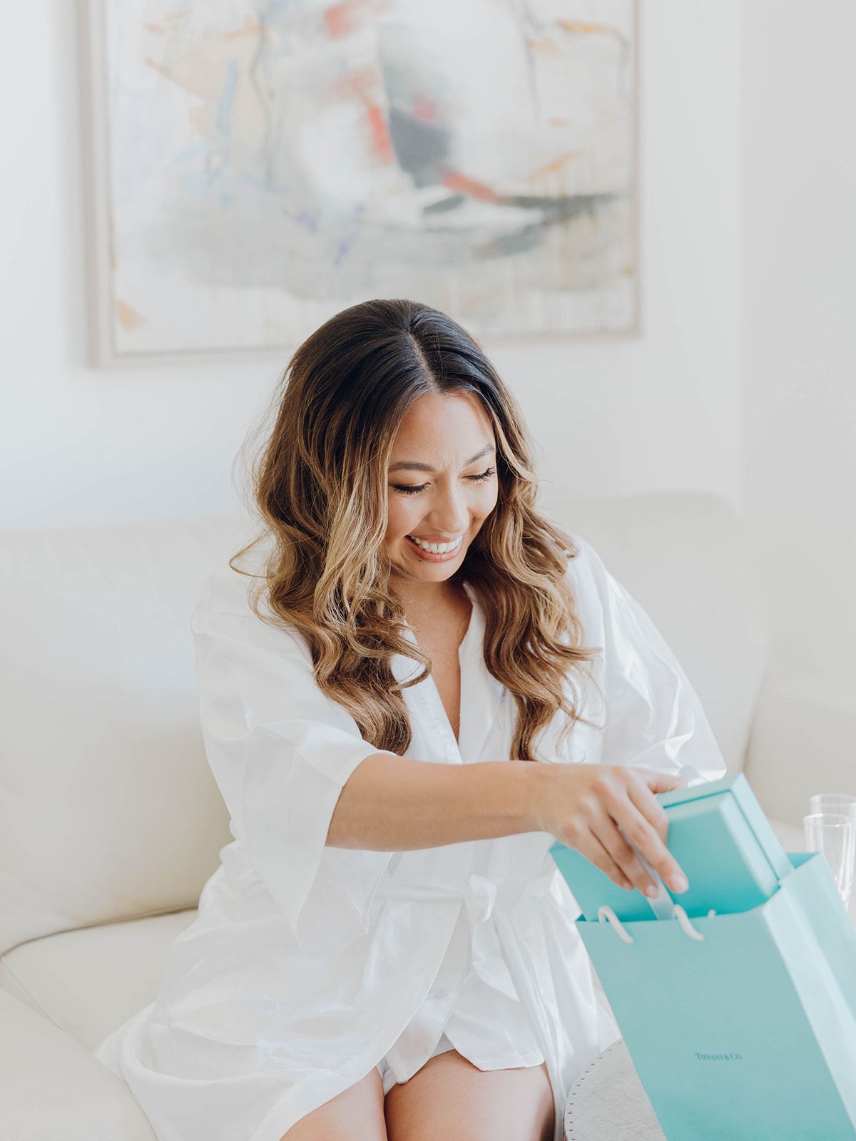 Woman in white robe opens a mint green gift bag while sitting on white couch with abstract art in background.