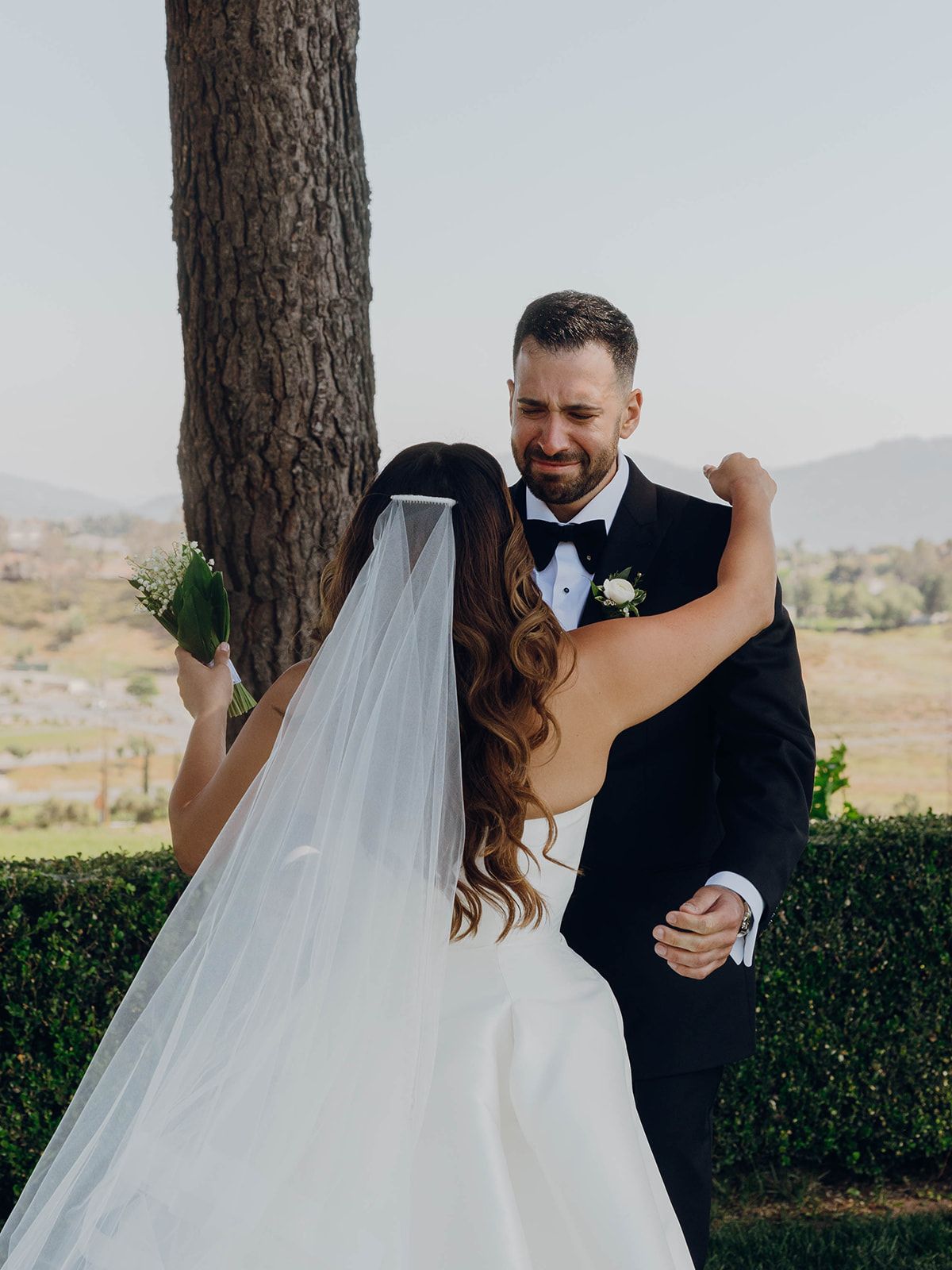 A bride and groom share their first dance under a tree during an outdoor wedding ceremony with scenic views.