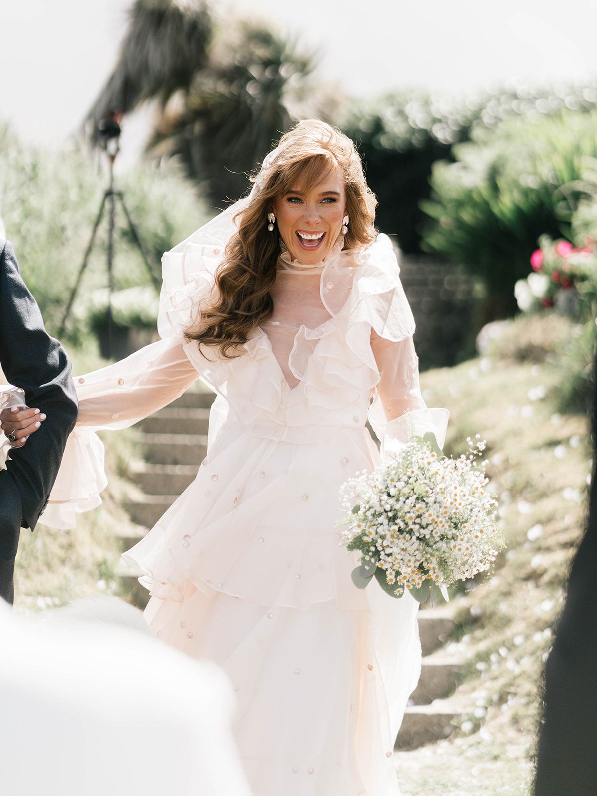 A joyful bride in a flowing white dress laughs while holding a bouquet of baby's breath flowers.