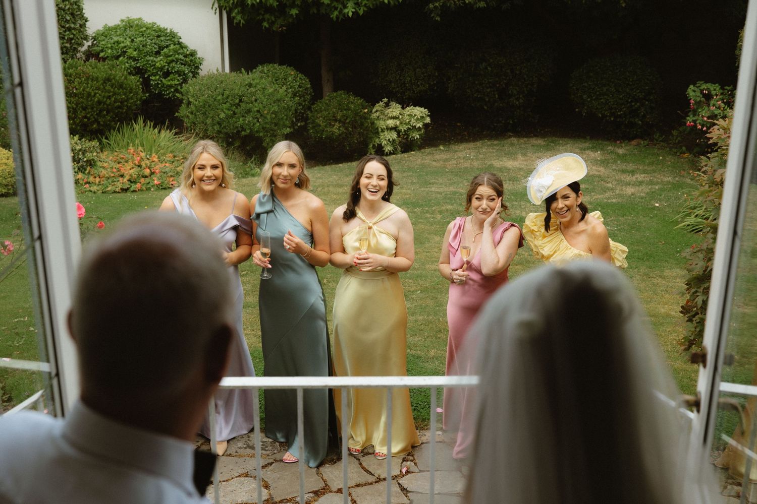 Wedding guests in pastel dresses watching through glass doors with drinks in hand.