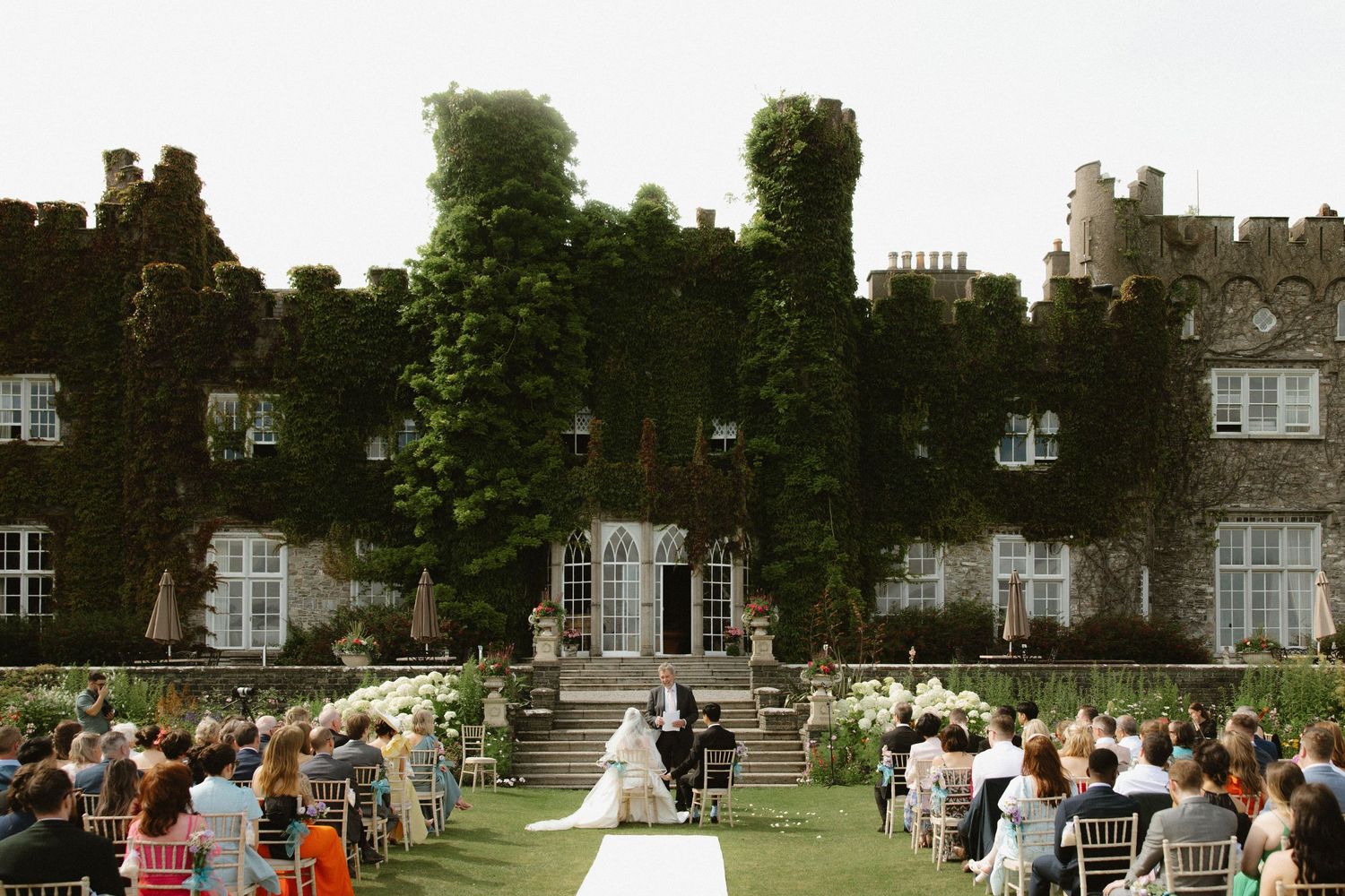 Wedding ceremony outside a grand ivy-covered stone castle with Gothic architecture.