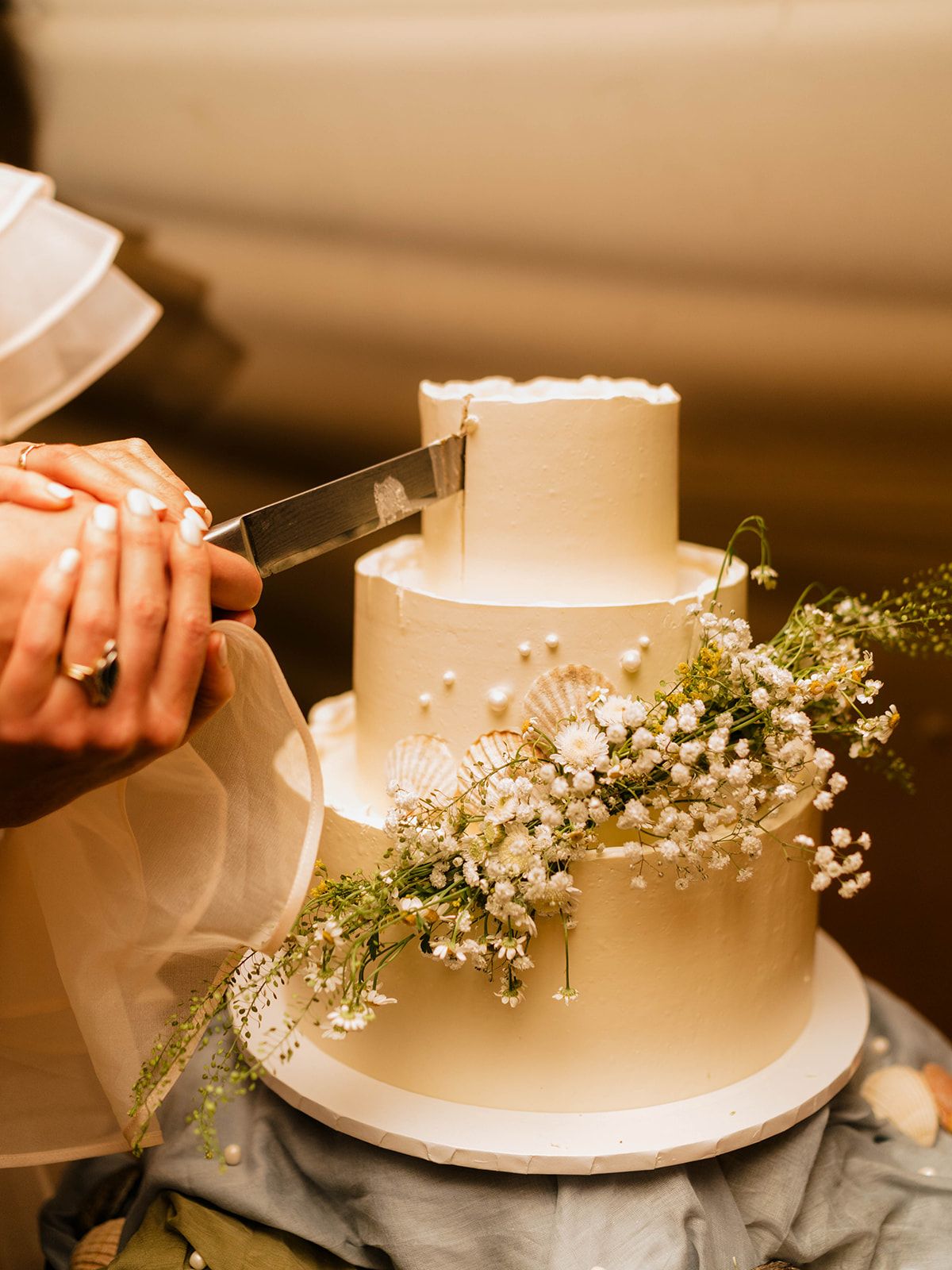 Close up of hands cutting into an elegant three-tier white wedding cake decorated with baby's breath.