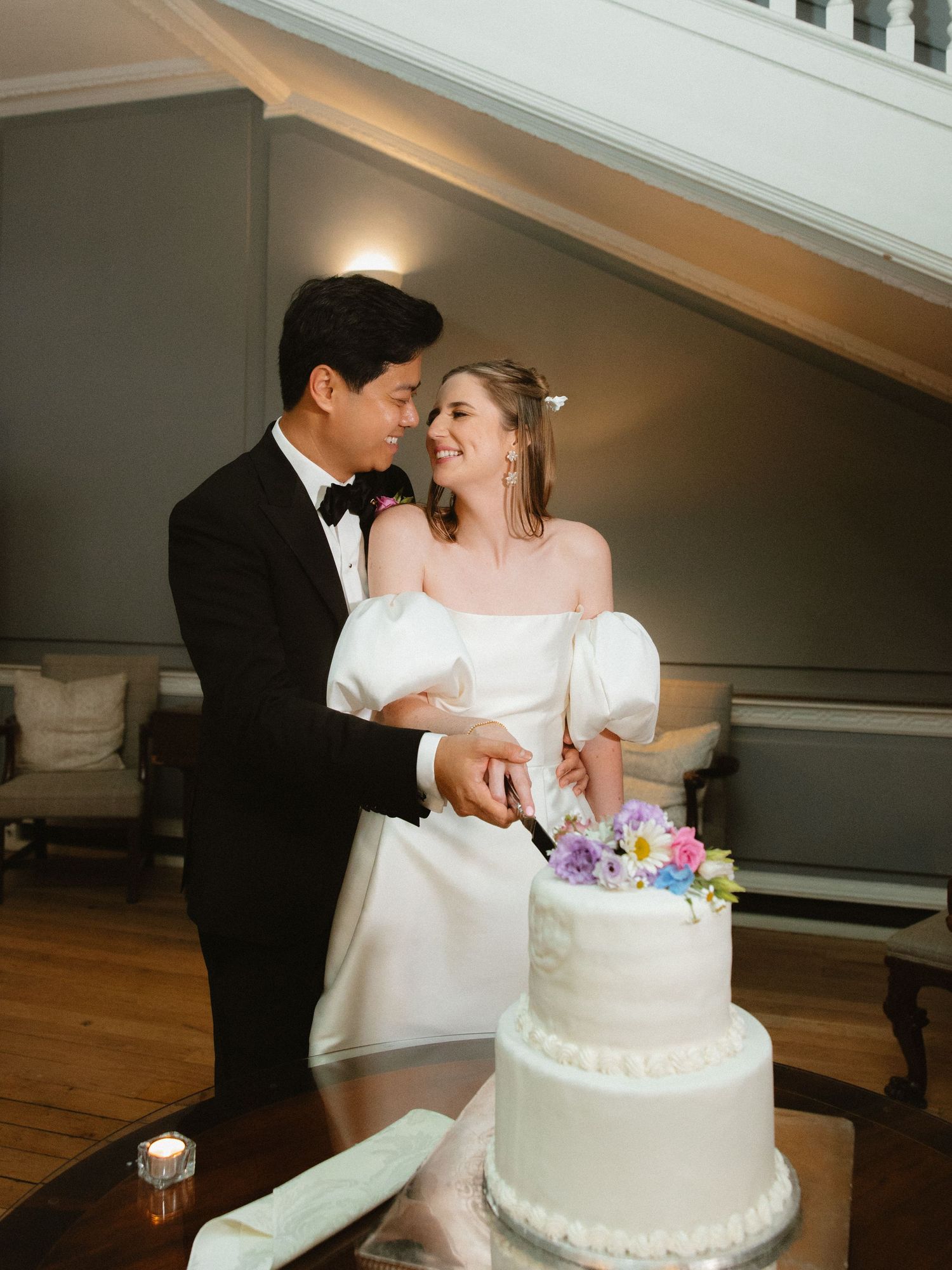 A couple in wedding attire smiling while cutting their white tiered wedding cake together.