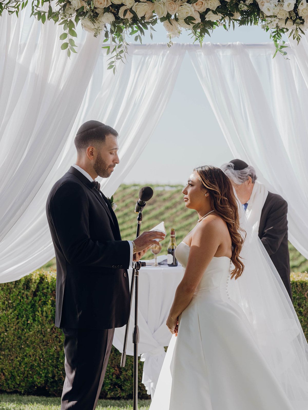 A couple exchanges vows under a white draped canopy with floral decorations during an outdoor ceremony.
