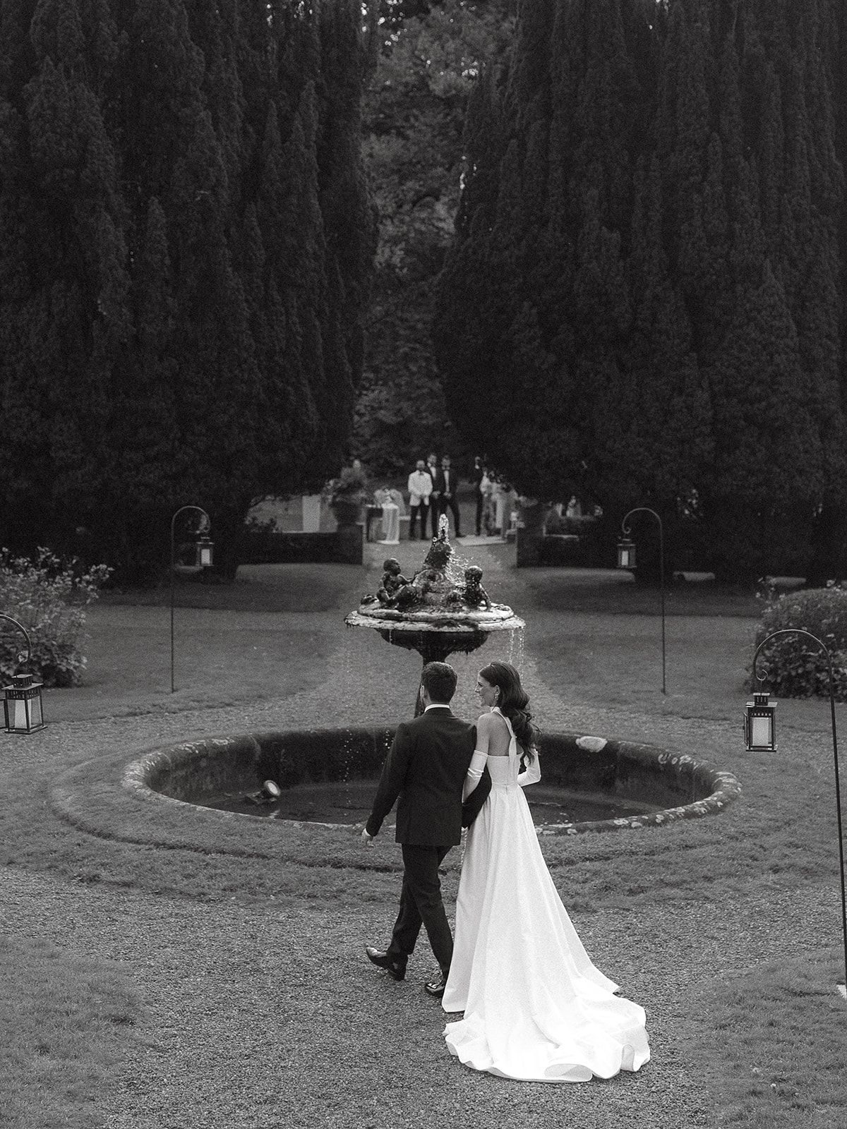 Wedding couple walks along a garden path lined with tall cypress trees.