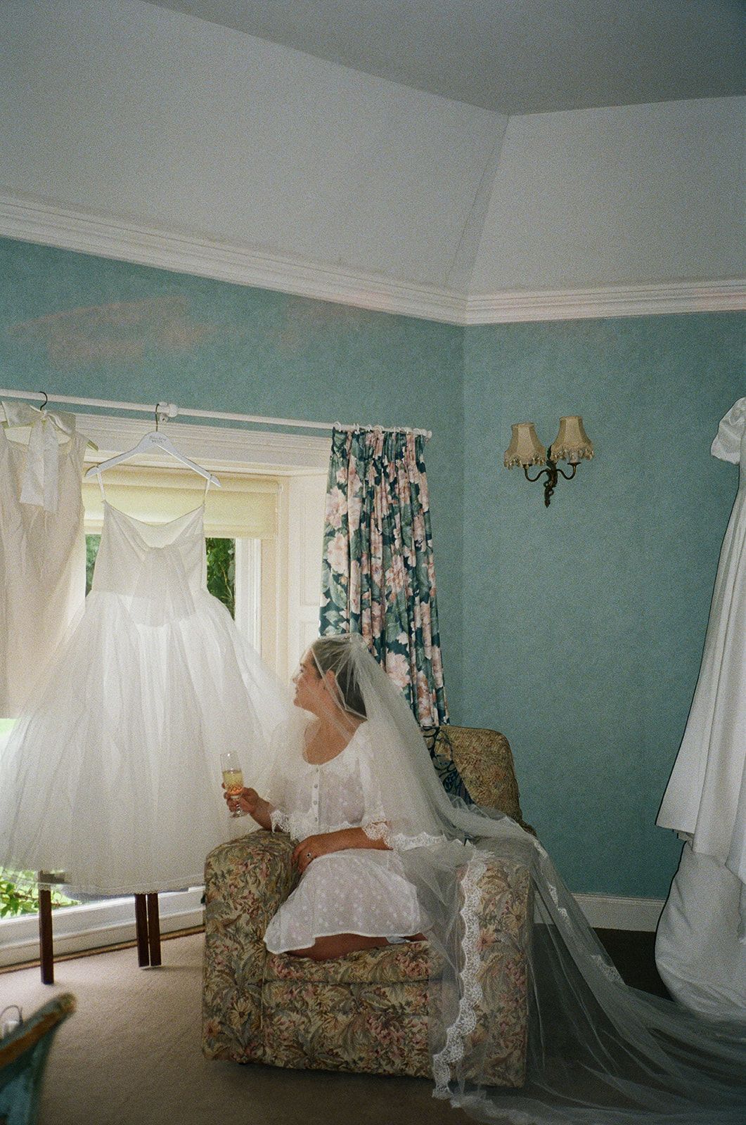 A white wedding gown hangs in a mint green room with floral curtains while someone sits in a floral armchair.