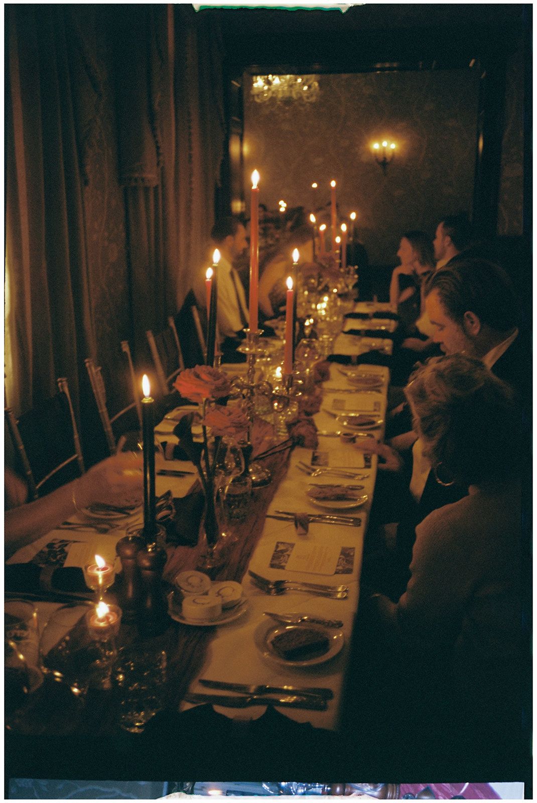 Candlelit dinner table set with white linens and glassware in a dimly lit room with multiple guests seated.