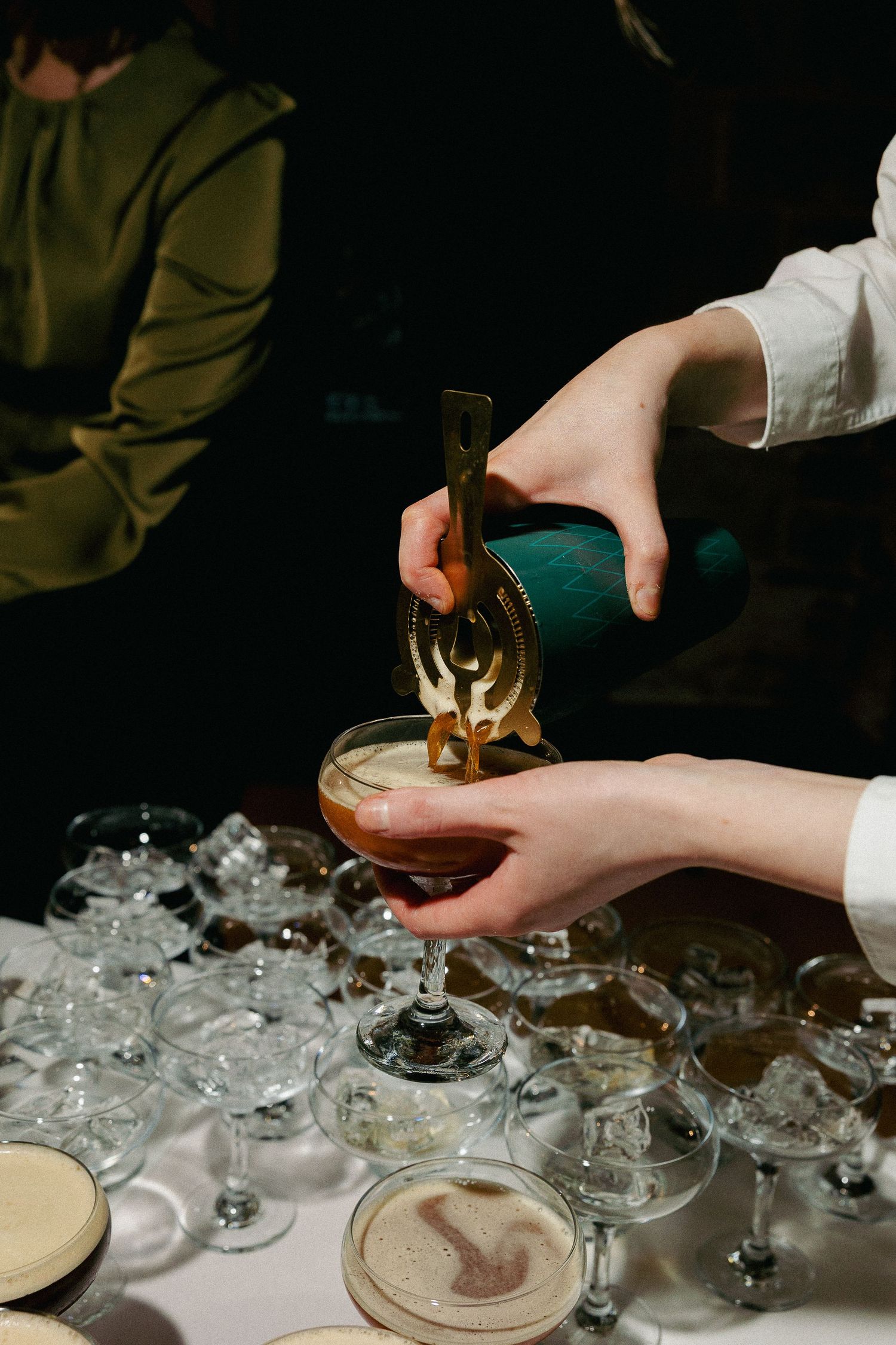 Hands pouring cocktails into multiple vintage coupe glasses arranged on a table with crushed ice.